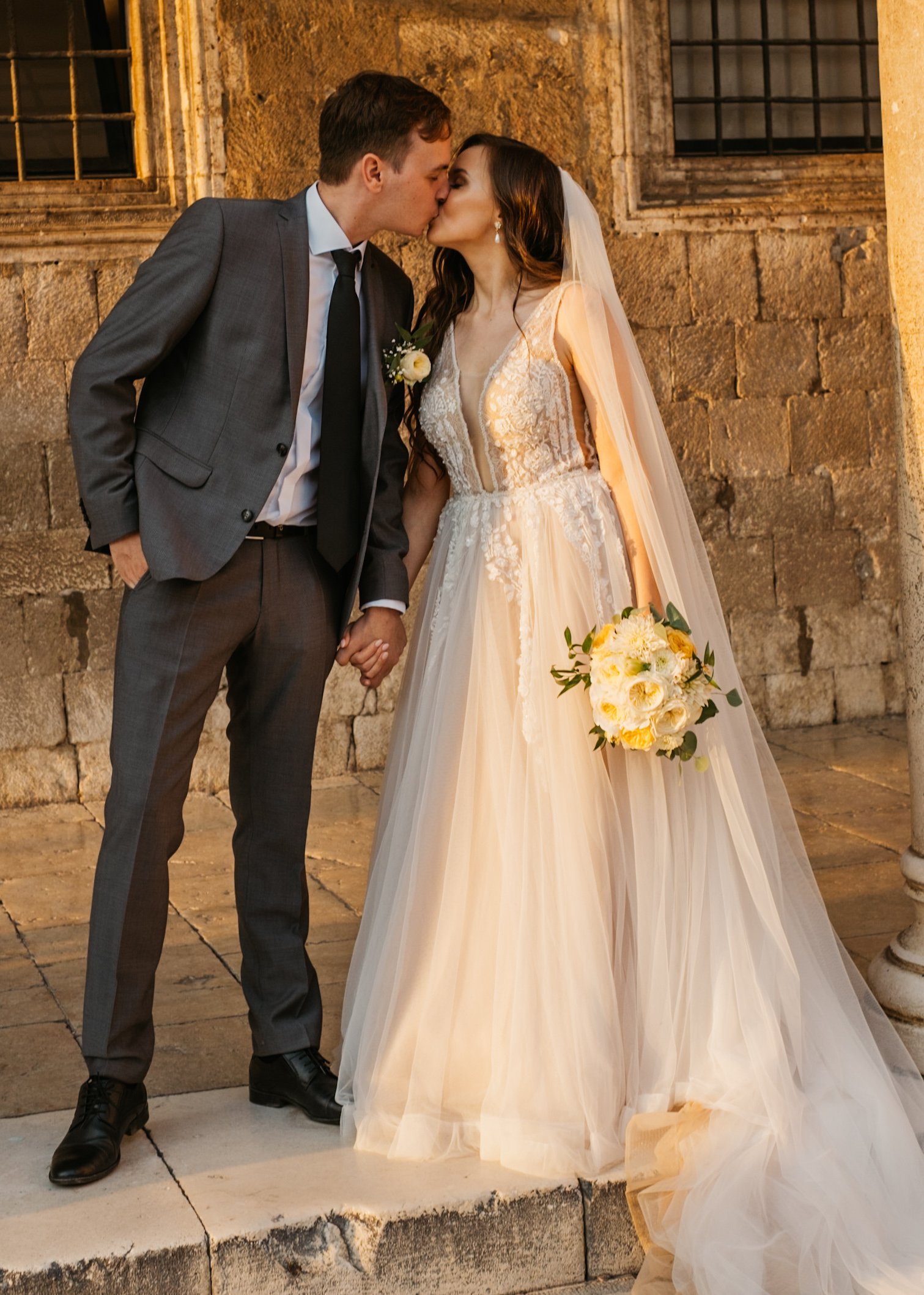 Bride and groom kissing outside a stone building, bride holding a bouquet.