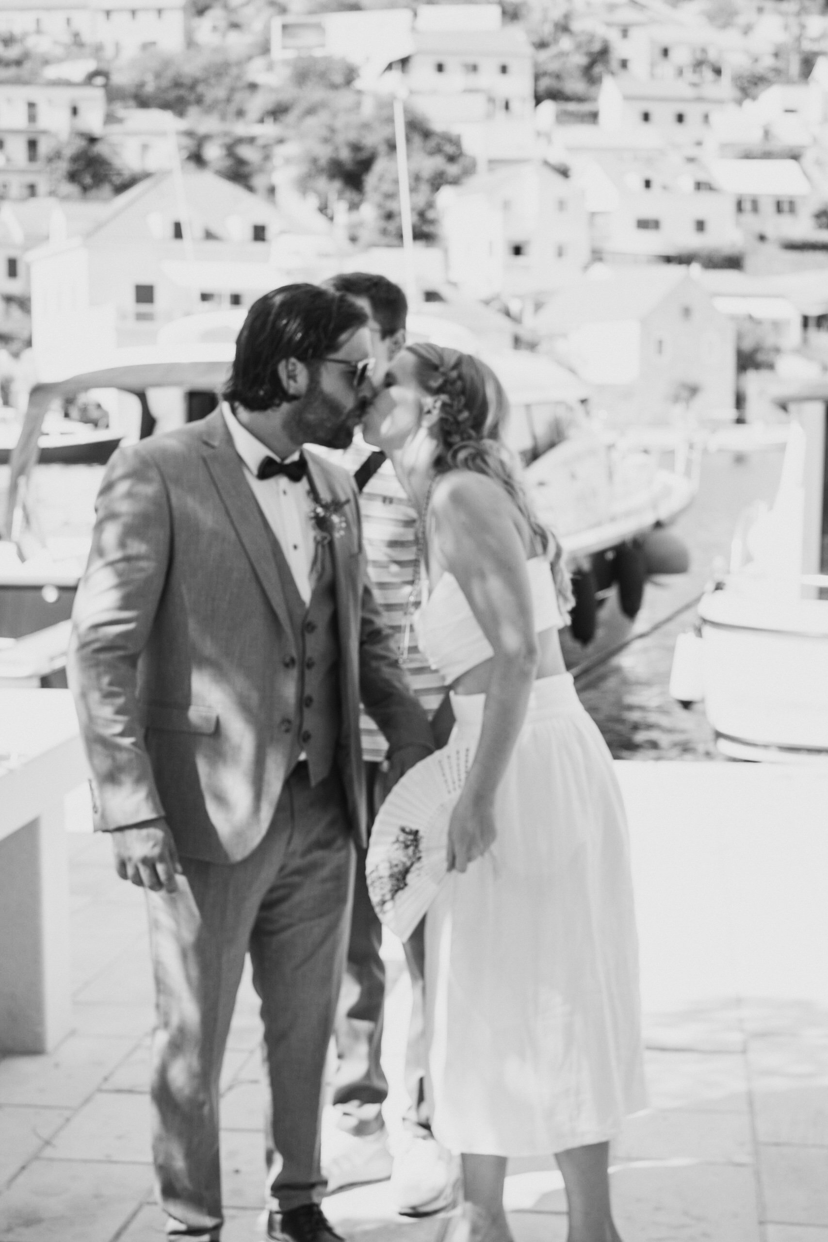 Bride and groom kissing at waterfront with boats and buildings in background, black and white photo.