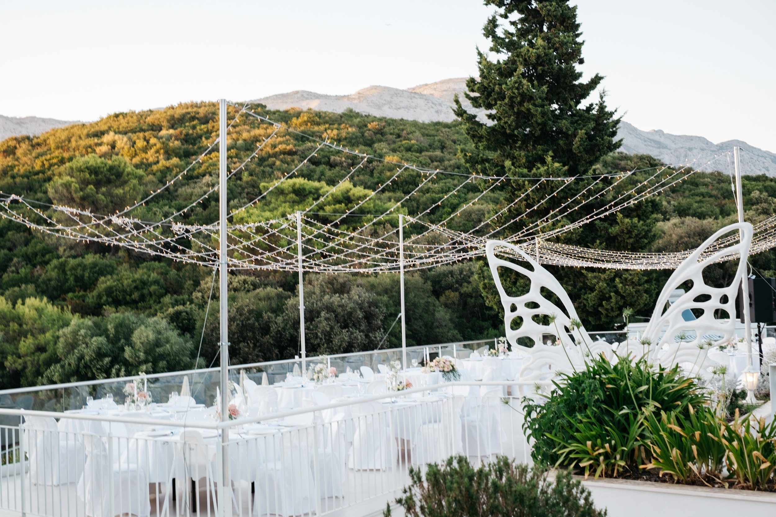 Outdoor wedding reception with hanging string lights, white tables and chairs, butterfly-shaped decor, and surrounding greenery at Korcula island in Croatia.