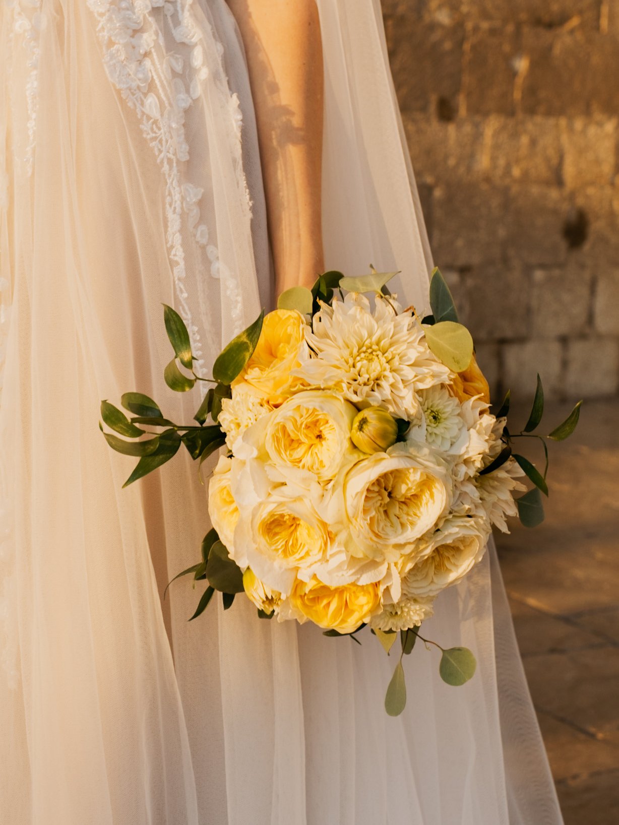 Bride holding a bouquet of yellow and white flowers with greenery, wearing a lace wedding dress.