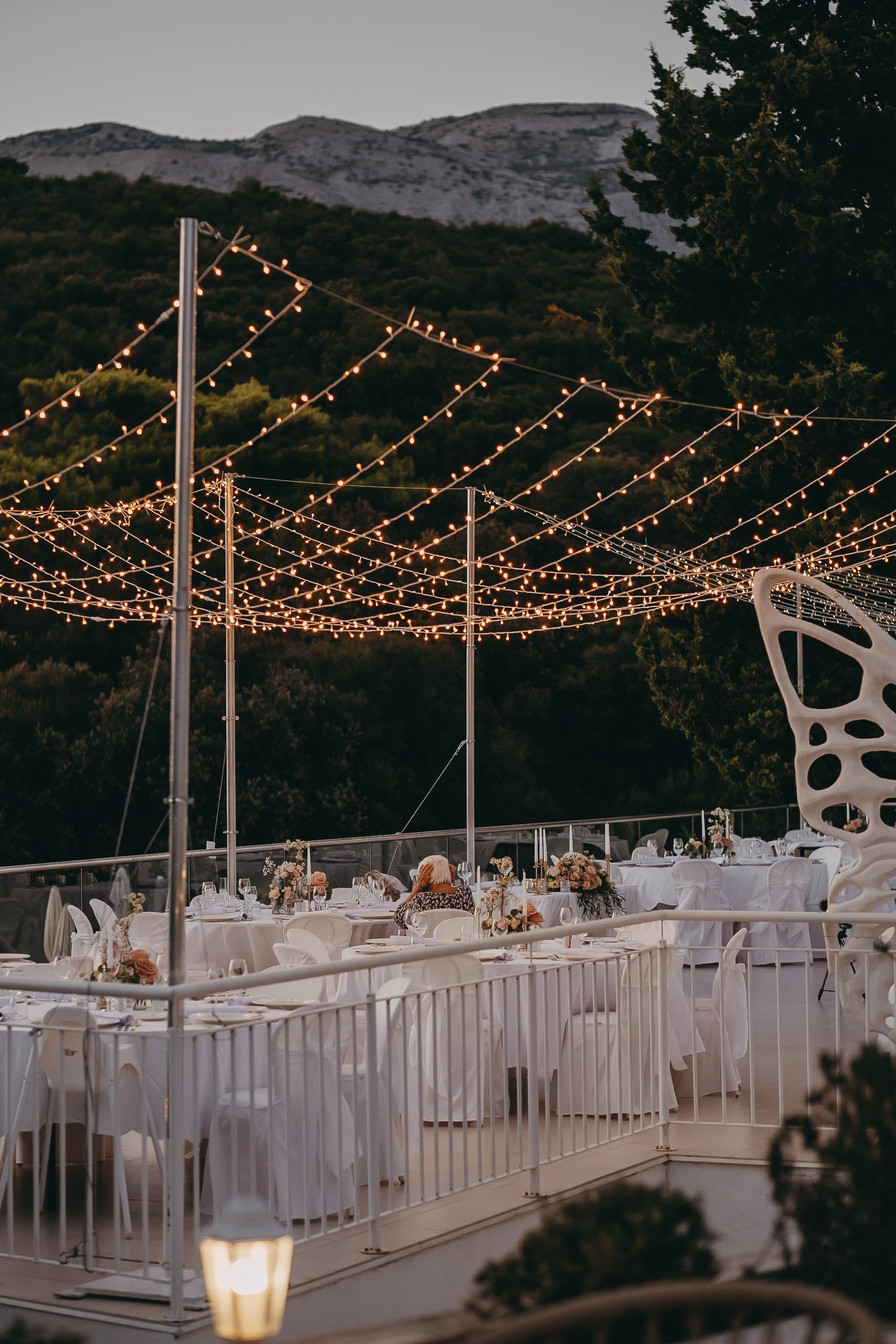 Outdoor wedding reception with string lights and decorated tables on a terrace, surrounded by mountains and trees at dusk.