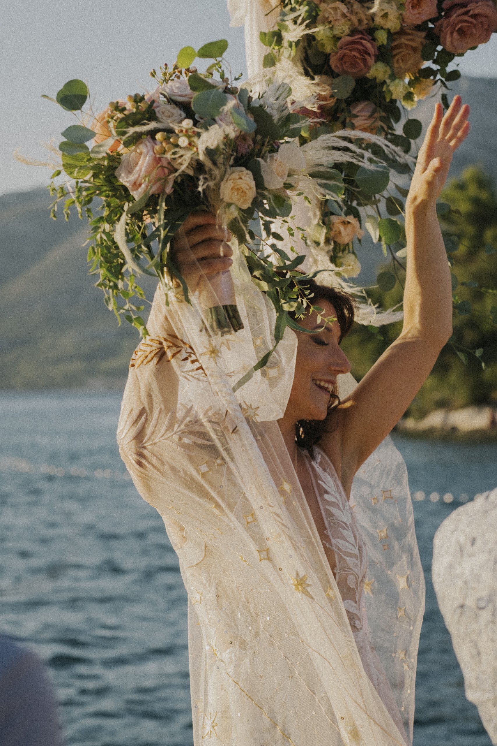 Bride holding a bouquet of flowers by a scenic water backdrop, wearing a decorative veil.