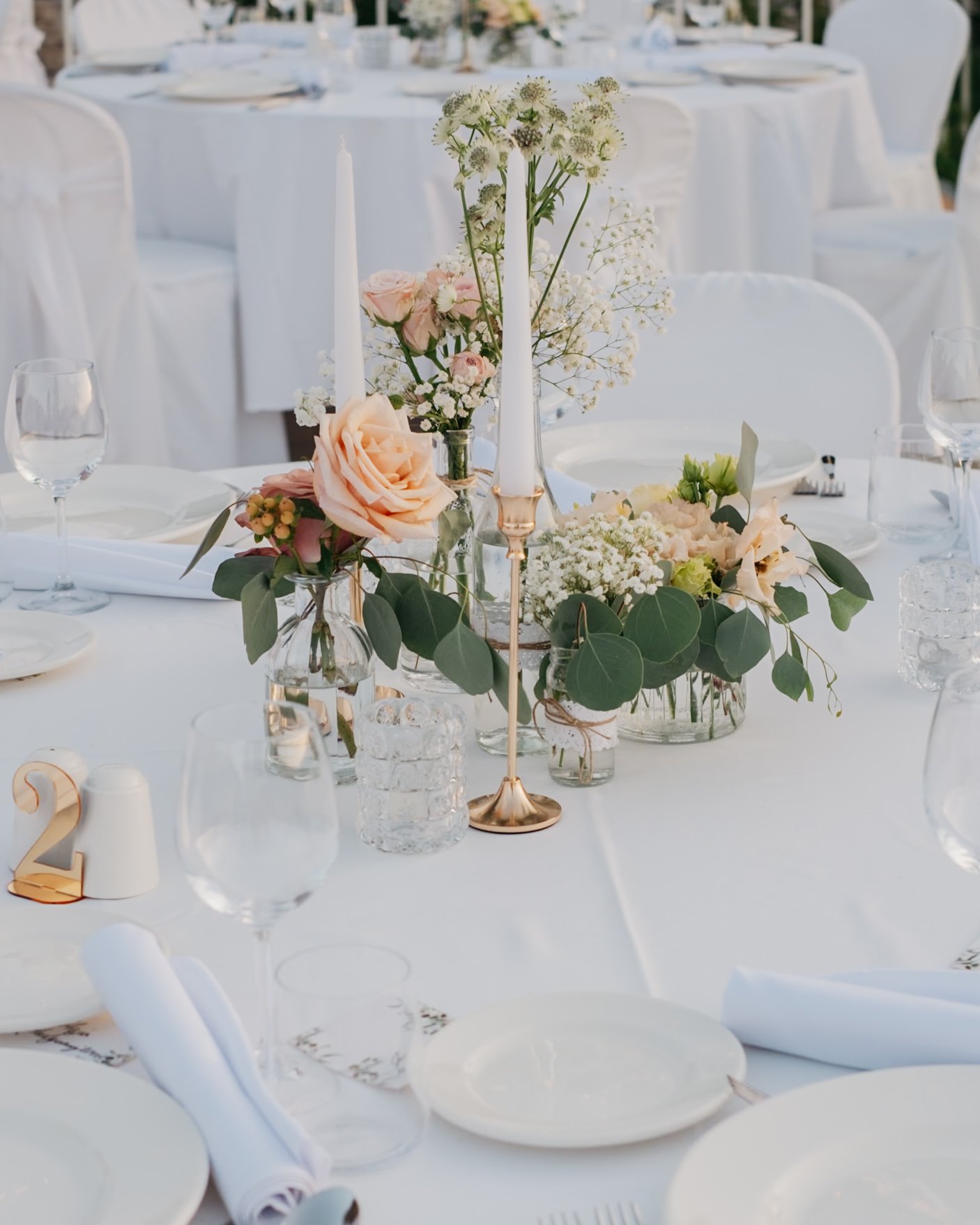 Elegant table setting with white tablecloth, floral centerpieces with roses and greenery, gold candle holders with white candles, plates, glassware, and table number two.