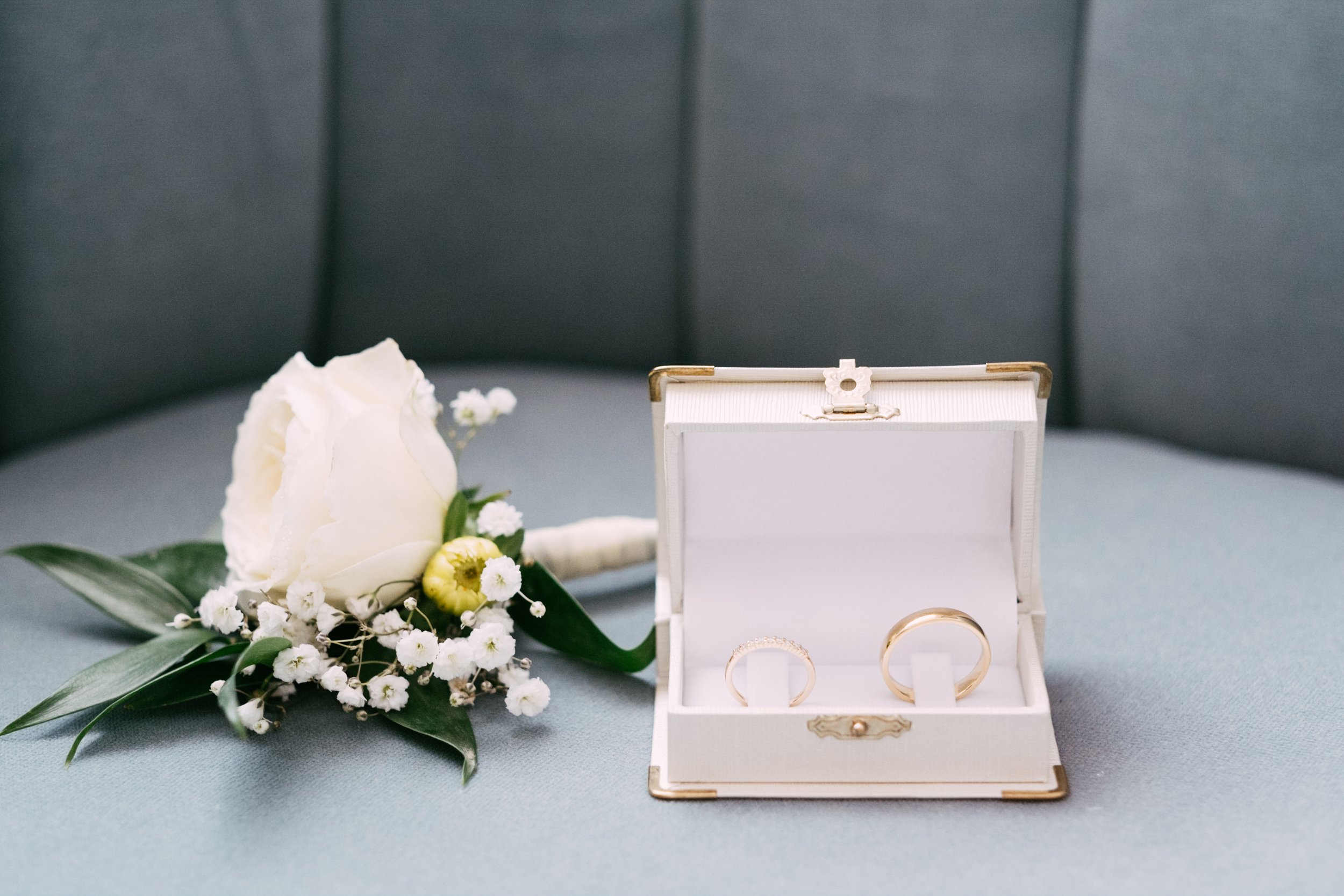 White rose boutonniere and wedding rings in an open box on a blue chair.