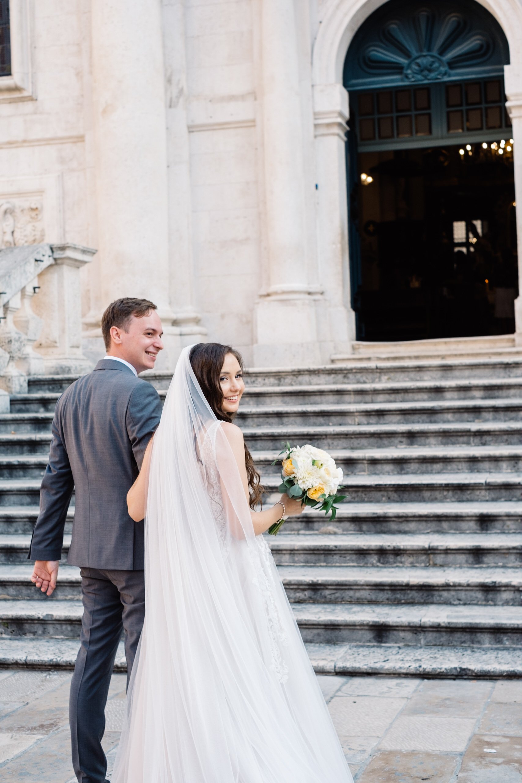 Bride and groom holding hands on stone steps, woman in wedding dress with veil and bouquet in front of Saint Blaise church in Dubrovnik.