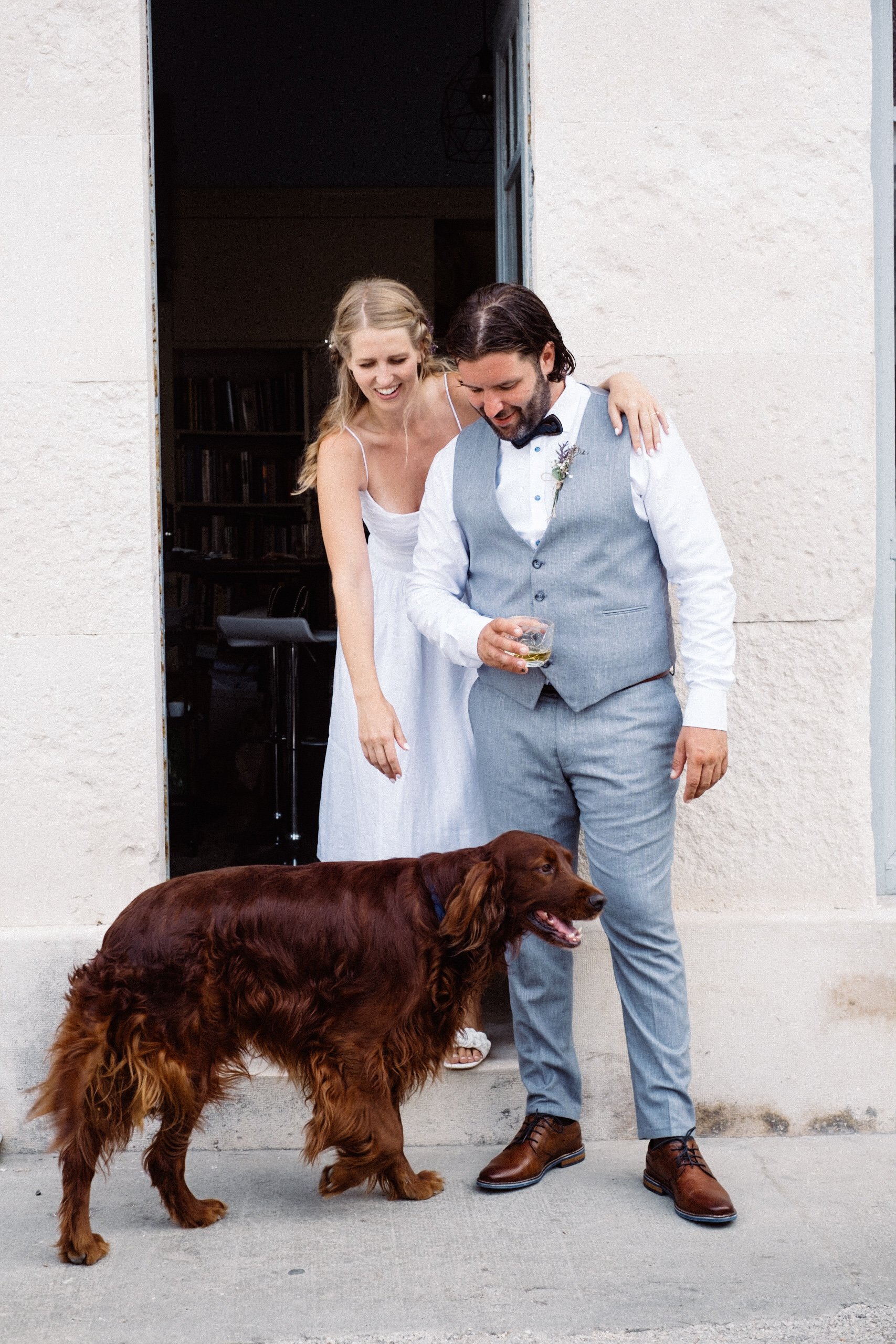 Bride and groom with Irish Setter dog standing outside a building, groom in grey suit and bride in white dress.
