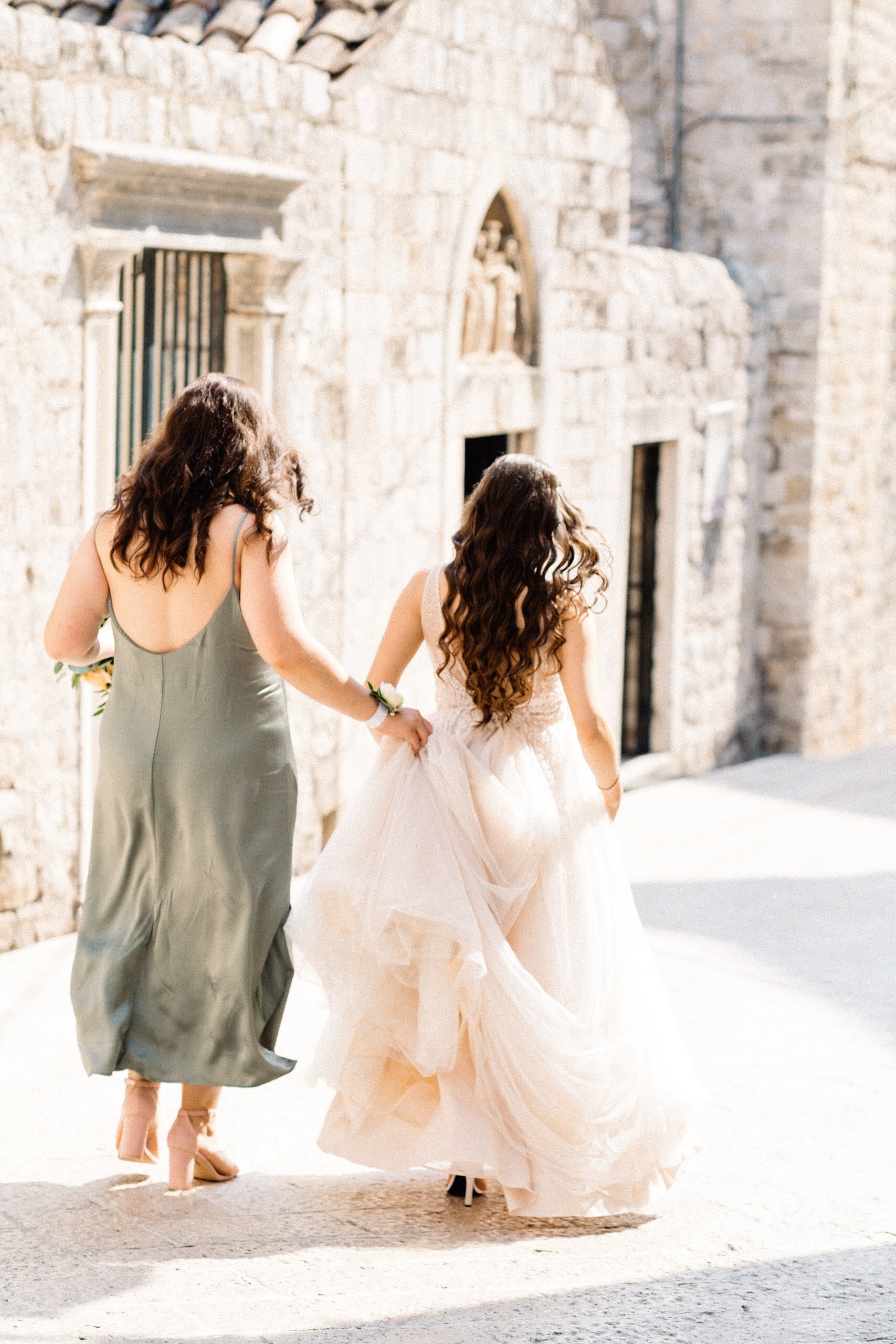 Two women walking in elegant dresses near a stone building, one in a sage green dress and the other in a wedding dress, hands gently holding the train.