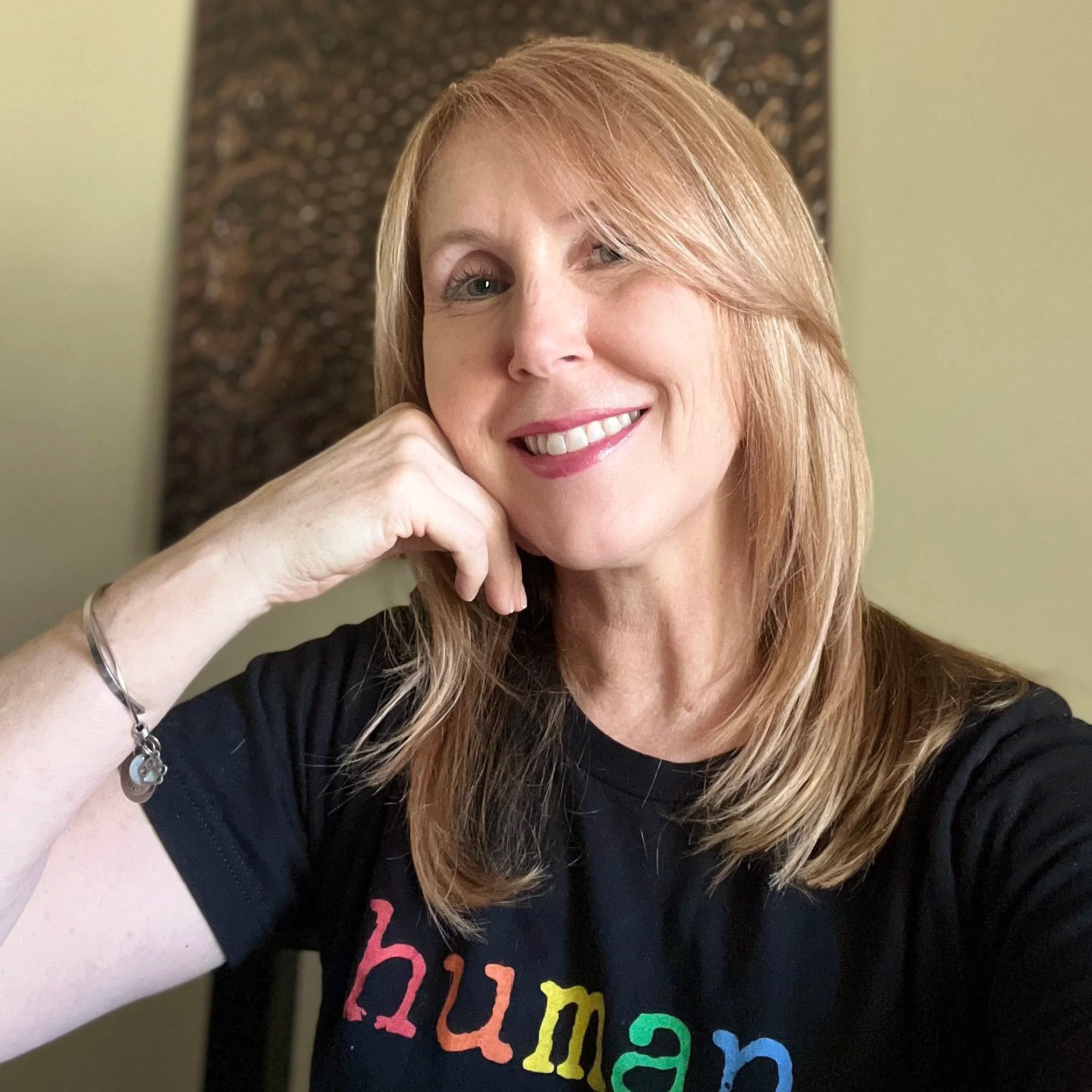 A woman with blonde hair smiling, holding her chin with her right hand, wearing a black shirt with colorful text that says "dream" and some bracelet accessories.