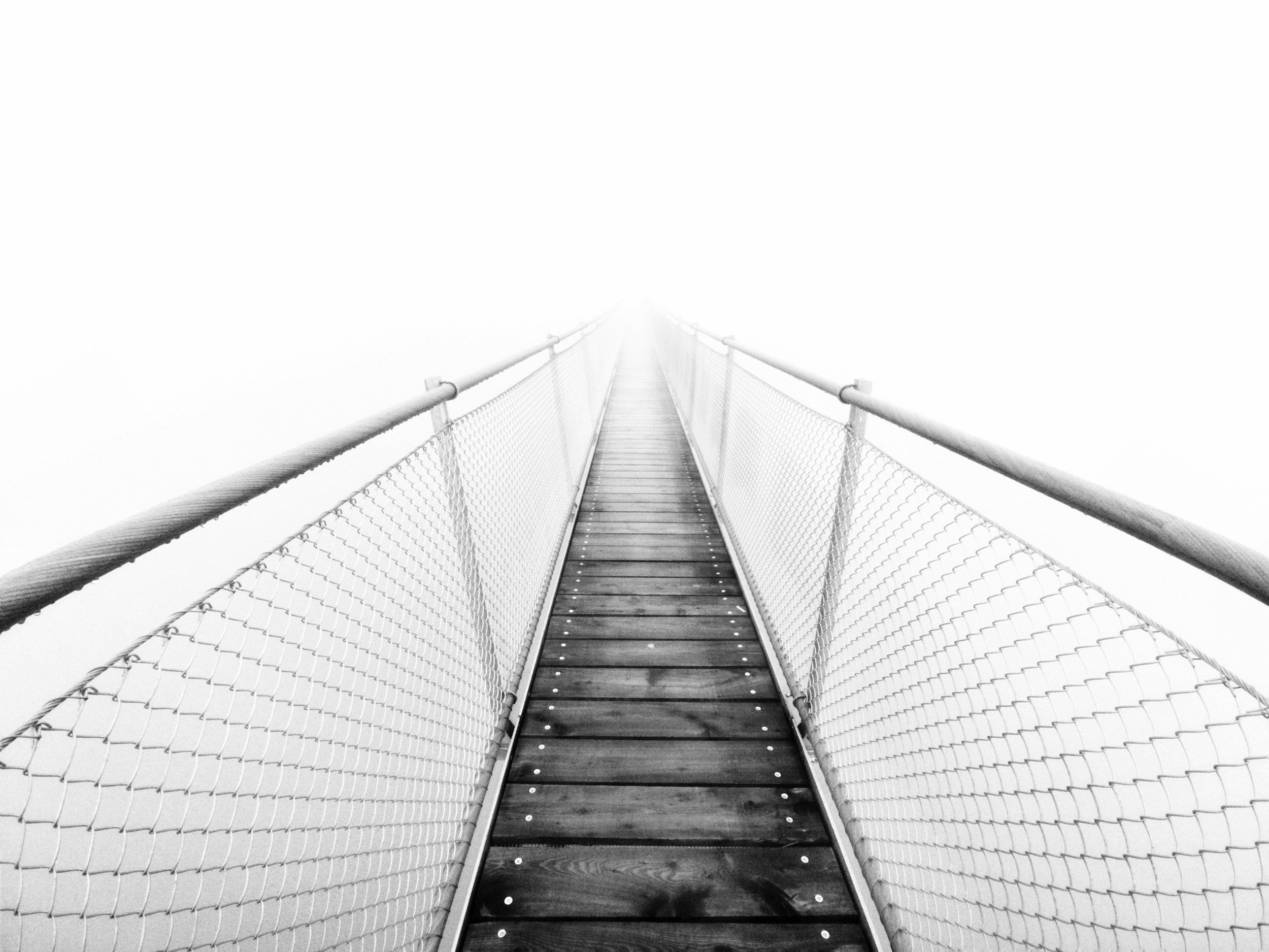 A narrow suspension bridge with wooden planks and chain-link sides extending into a bright, white foggy background, creating an illusion of endlessness and mystery.