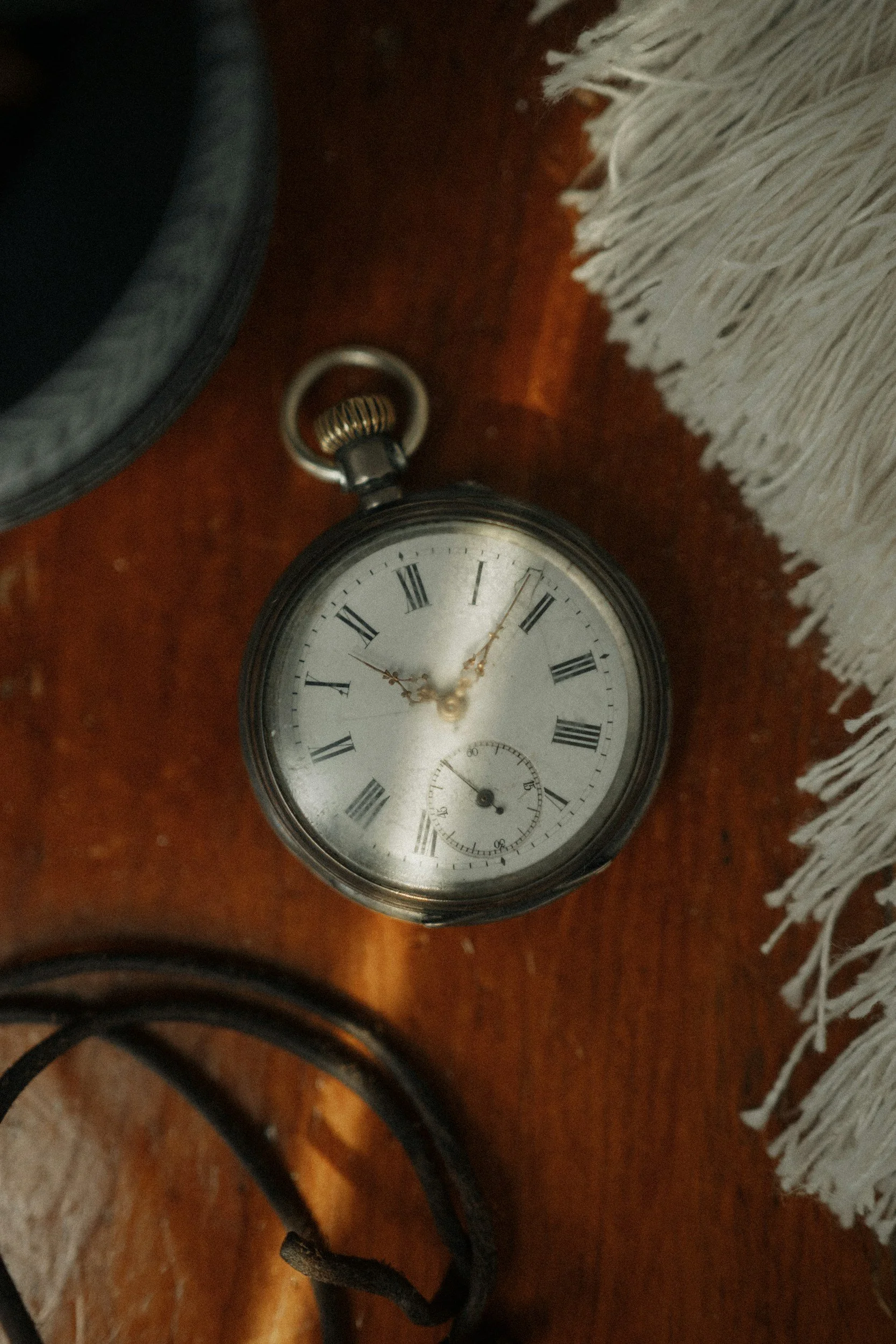 A vintage pocket watch with Roman numerals on a wooden surface, next to a white tassel and some electrical cords.