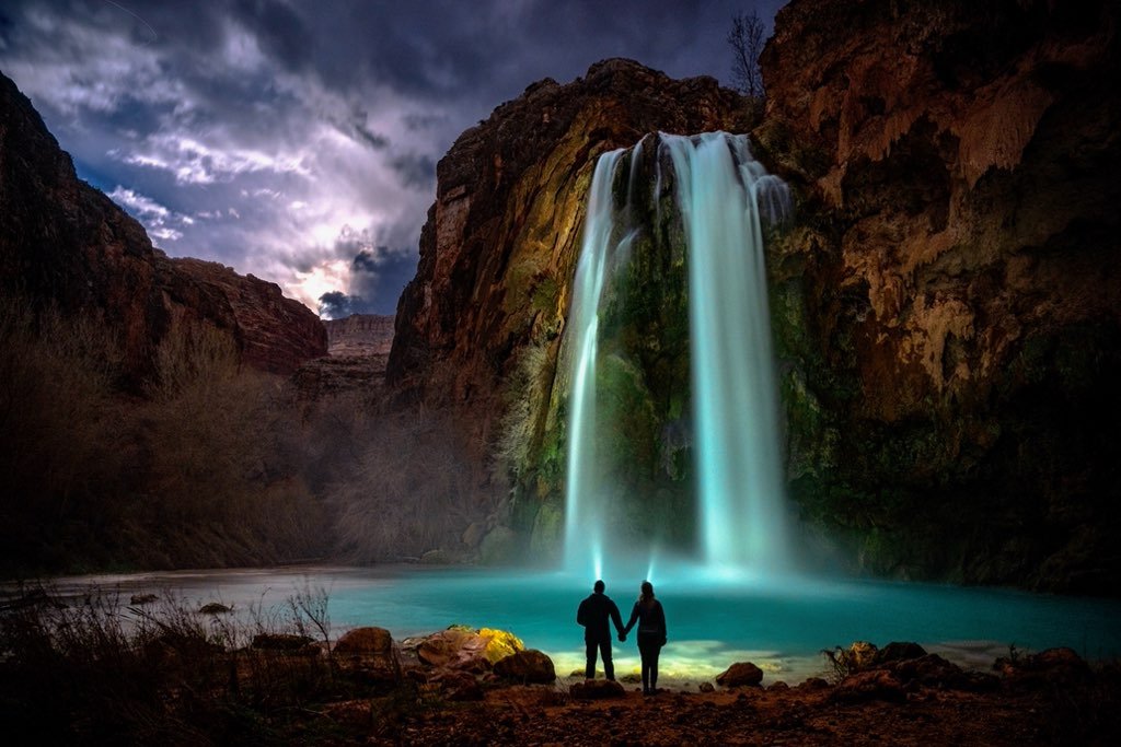 A couple holding hands and standing in front of a large waterfall at night.
