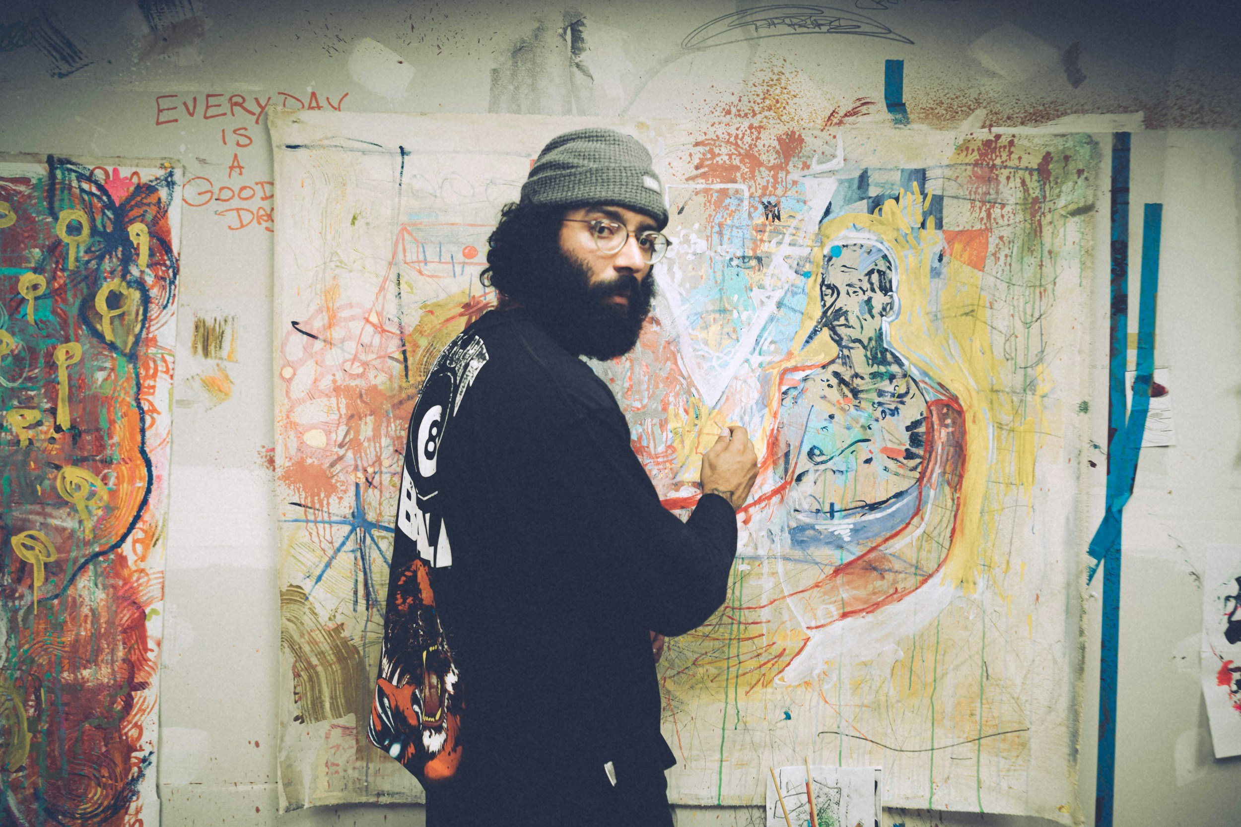 An artist with a beard, glasses, and a beanie hat working on a colorful abstract painting on a large canvas in his studio. The background features additional artworks and notes, including the phrase "EVERYDAY IS A GOOD DAY."