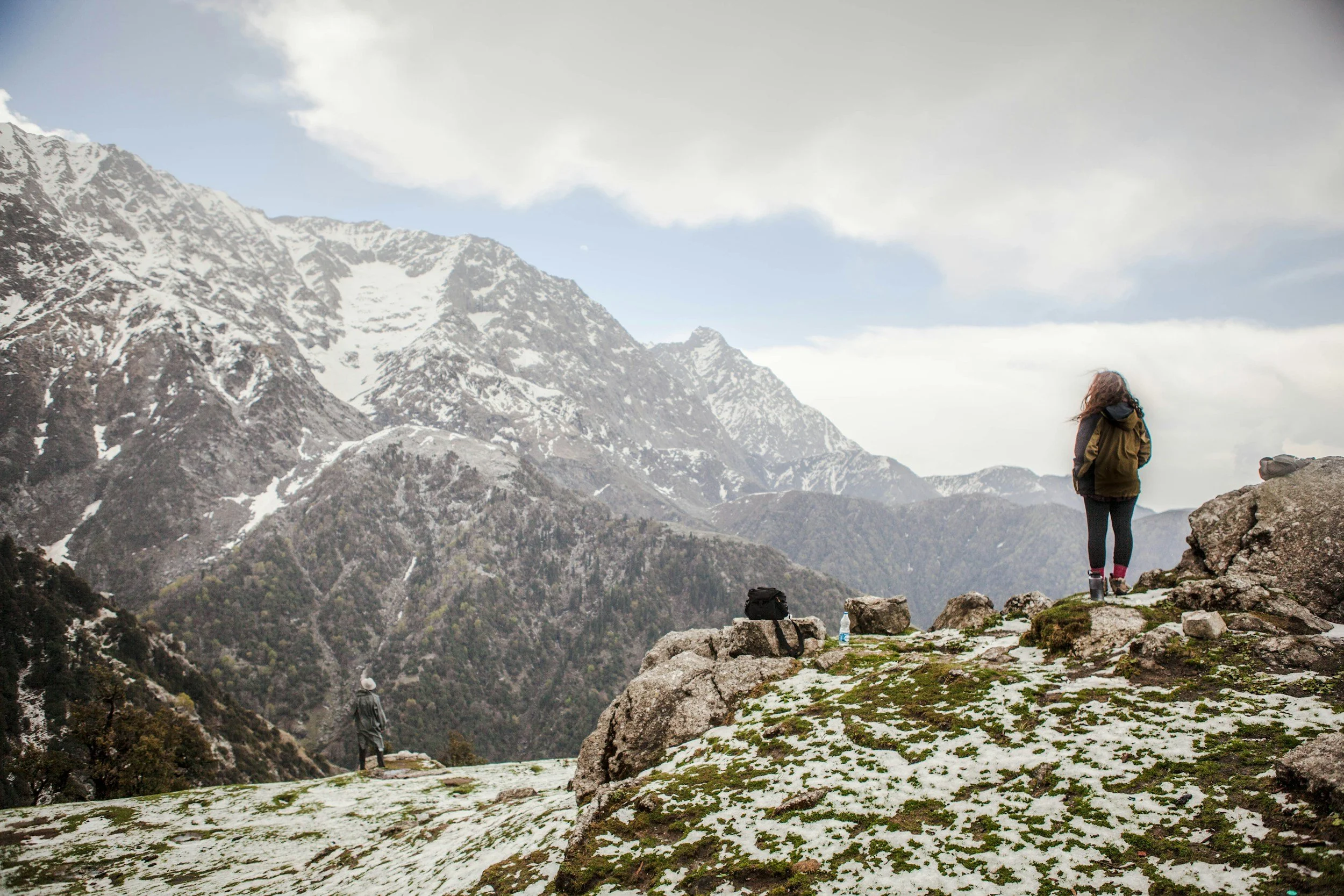 Two hikers standing on a rocky, mossy mountain ledge with snow patches, overlooking a mountain range with snow-capped peaks under a cloudy sky.