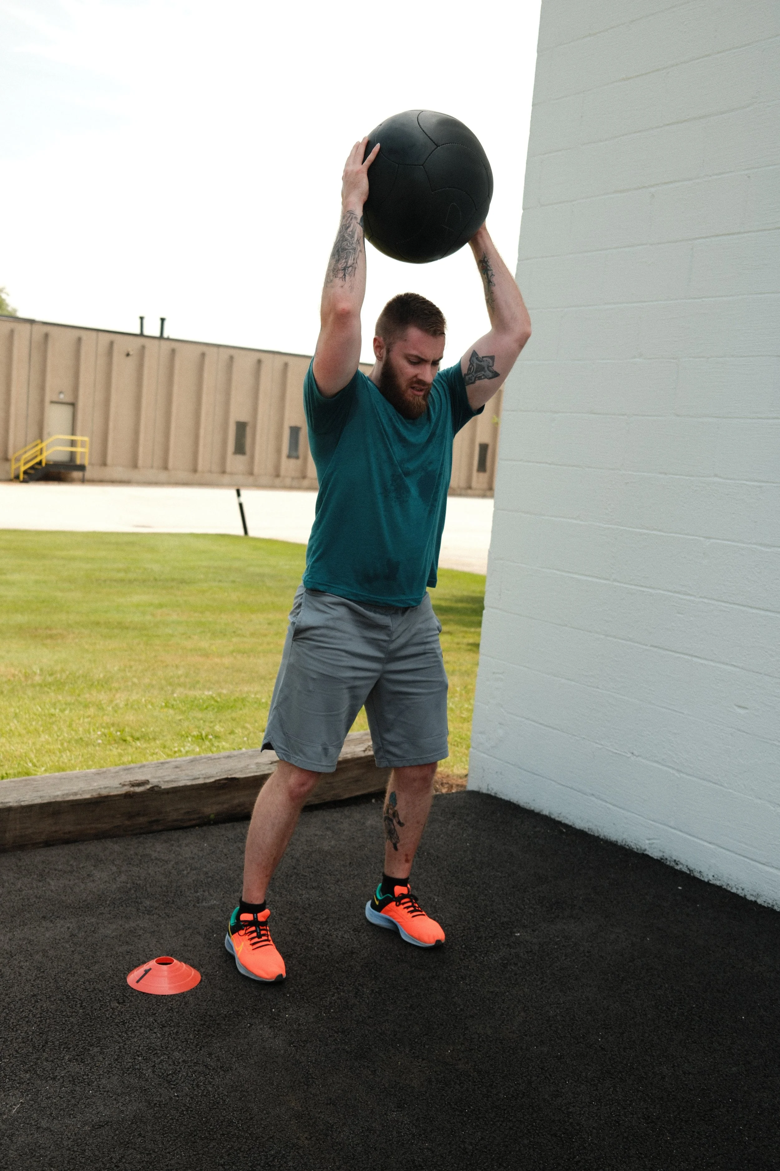 A man with a beard and tattoos, dressed in a teal shirt, gray shorts, and bright orange athletic shoes, is lifting a large black medicine ball above his head outside near a white wall, with a grassy area and a building in the background.