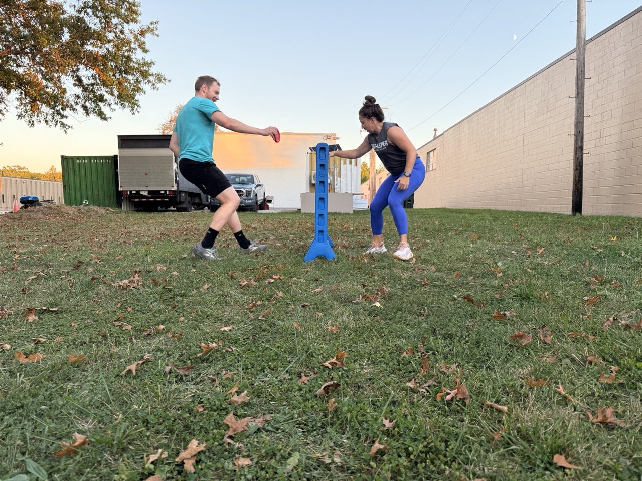 Two people playing a game of giant Connect Four outdoors on grass with fallen leaves, a large green storage container, a truck, a car, and a building in the background during late afternoon.