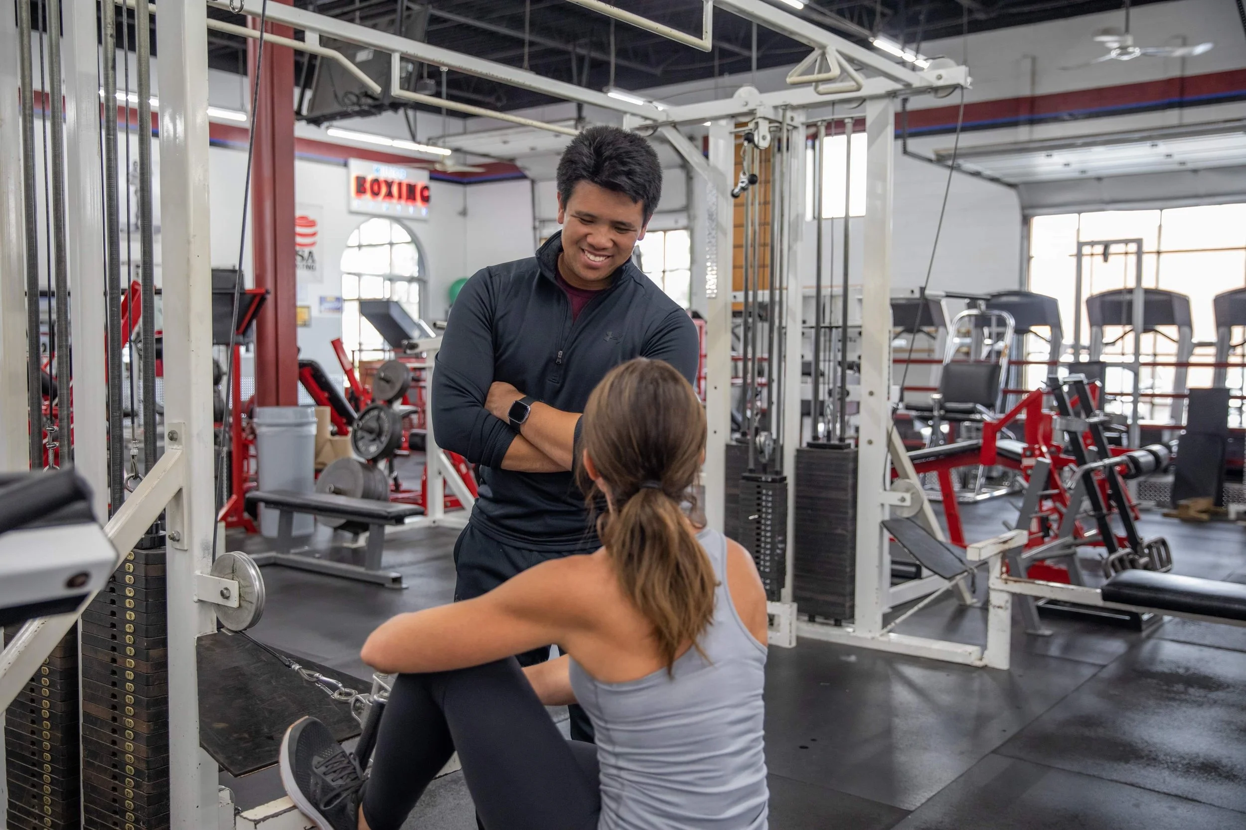 A man with short black hair and a smartwatch smiling and talking to a woman with long brown hair, who is seated on a bench at the gym. There are gym equipment and weights around them.
