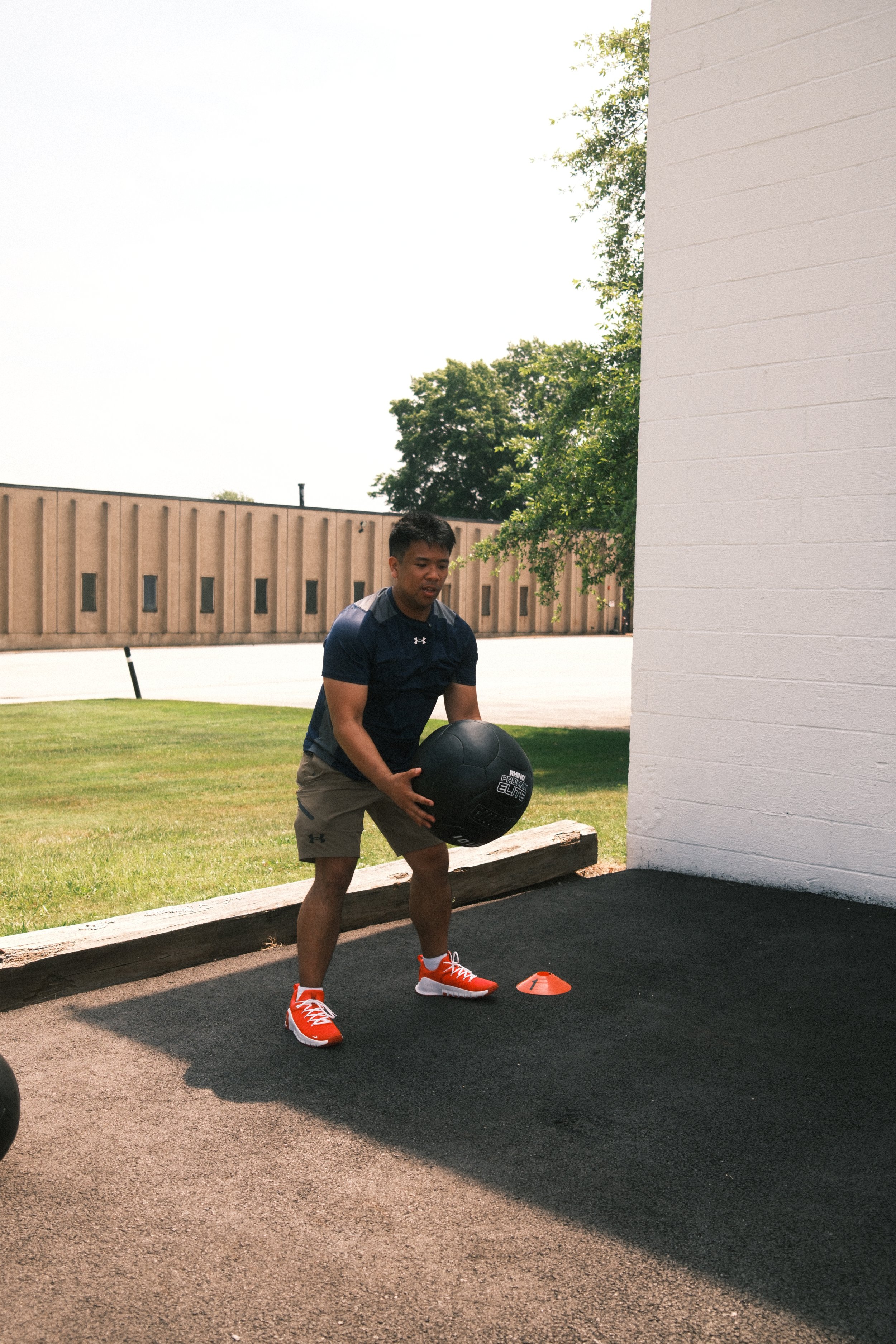 A man in athletic attire, red sneakers, and shorts holding a medicine ball during an outdoor workout session next to a white building wall.