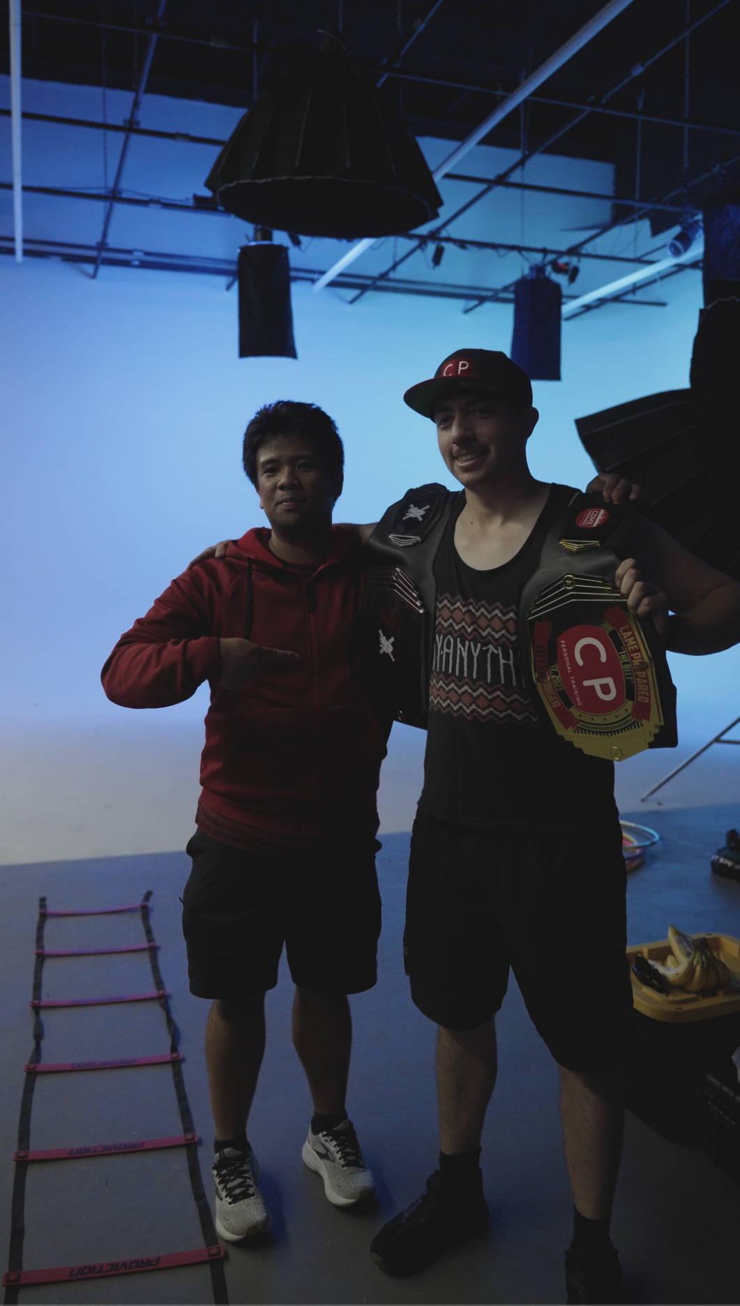 Two men standing together, one pointing at the other, inside a studio or gym. The man on the right is wearing a black cap, black shorts, and a WWE championship belt, while the man on the left is wearing a red hoodie and black shorts. Items like a pin