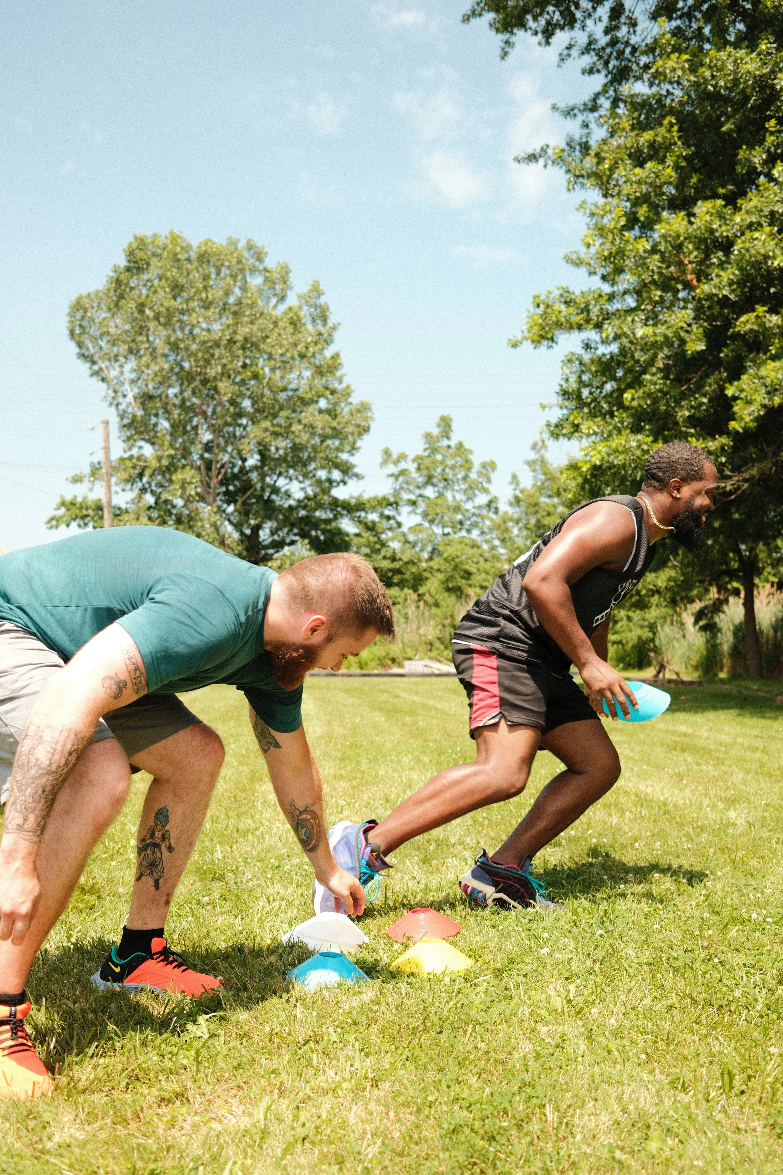 Two men preparing to start a race outdoors, crouched near colorful cones on grass with trees and blue sky in the background.