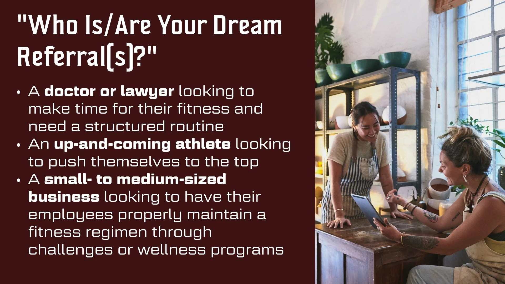 A woman smiling and talking to a tattooed woman sitting at a counter in a bright, industrial-style kitchen with shelves and plants, while holding a tablet.