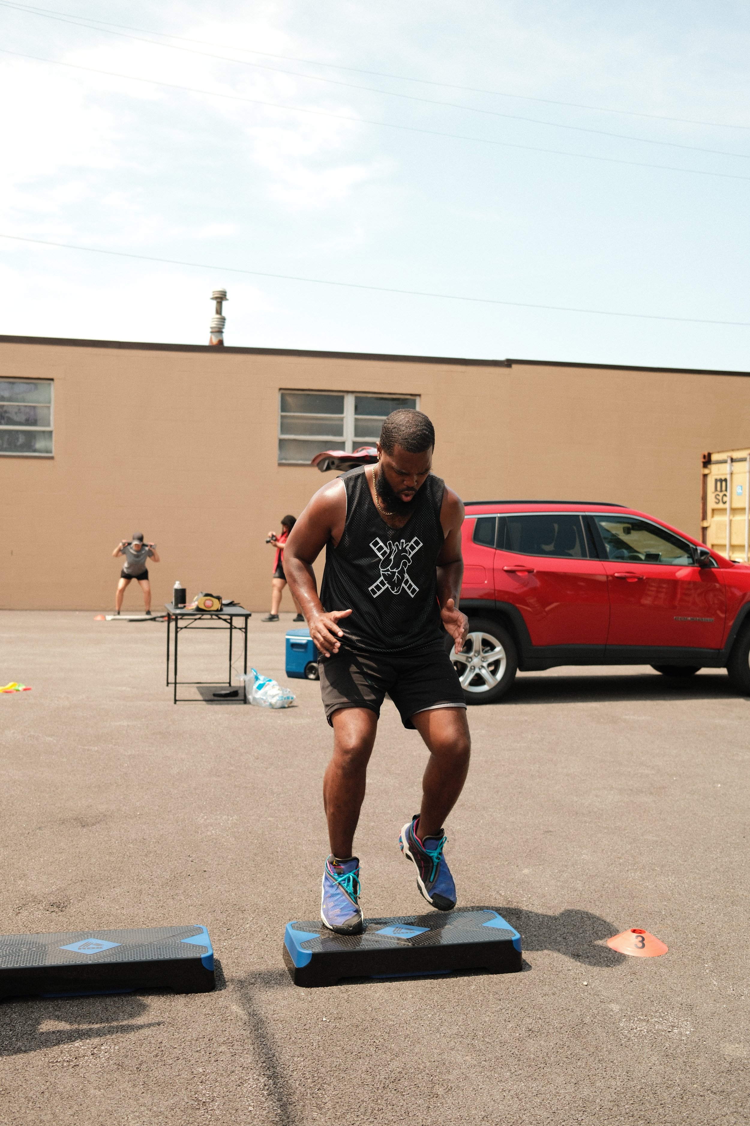 A man performs a step exercise on a balance board outdoors during a fitness event, with people and a red vehicle in the background.