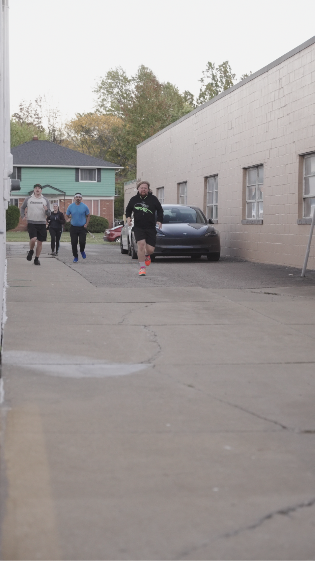 Four people are running on a sidewalk next to a building, with two cars parked nearby and trees in the background.