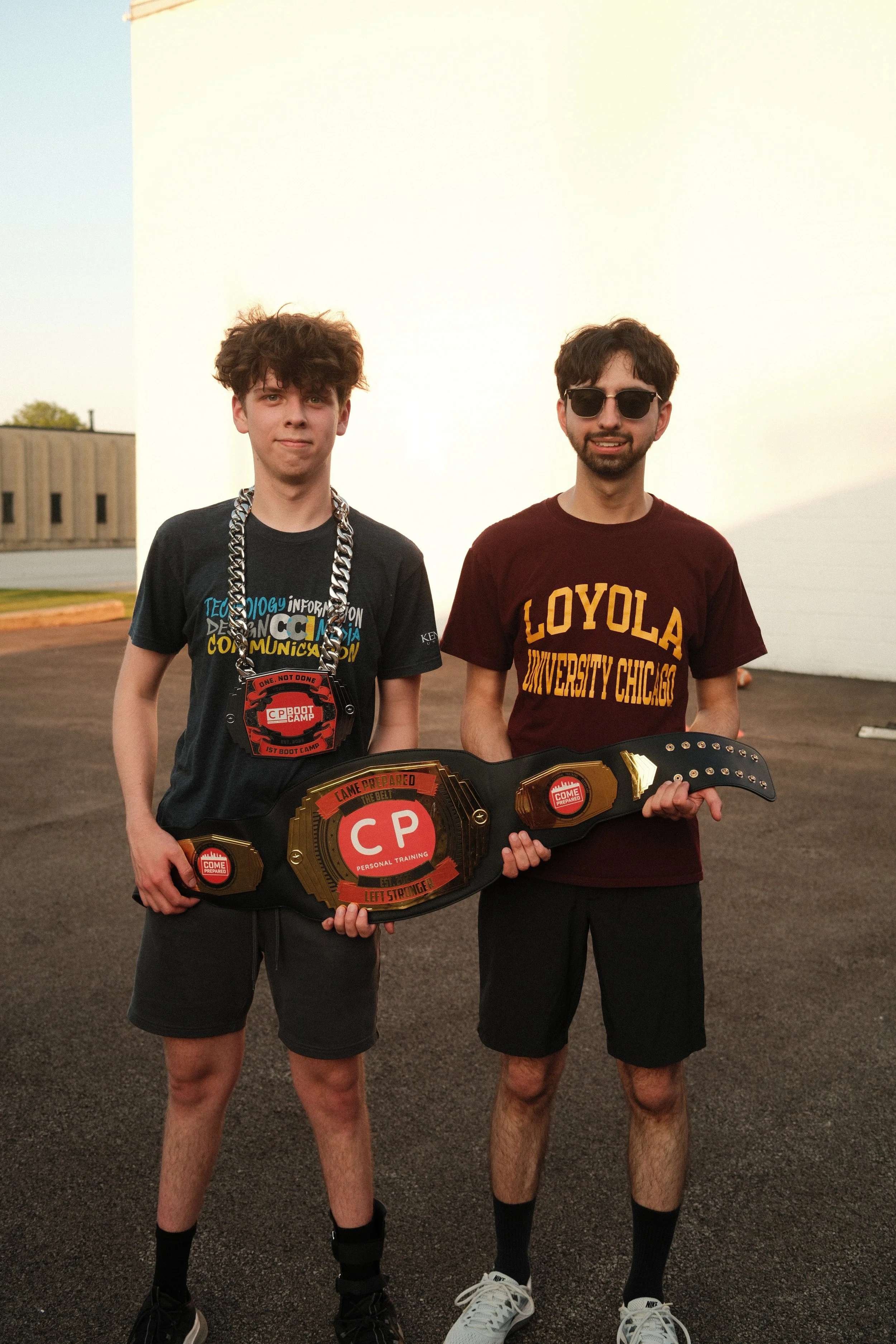 Two young men standing outdoors on a dark asphalt surface, holding a large championship belt with red and gold accents, with a white building and clear sky in the background. One is wearing sunglasses and a maroon Loyola University Chicago t-shirt, t