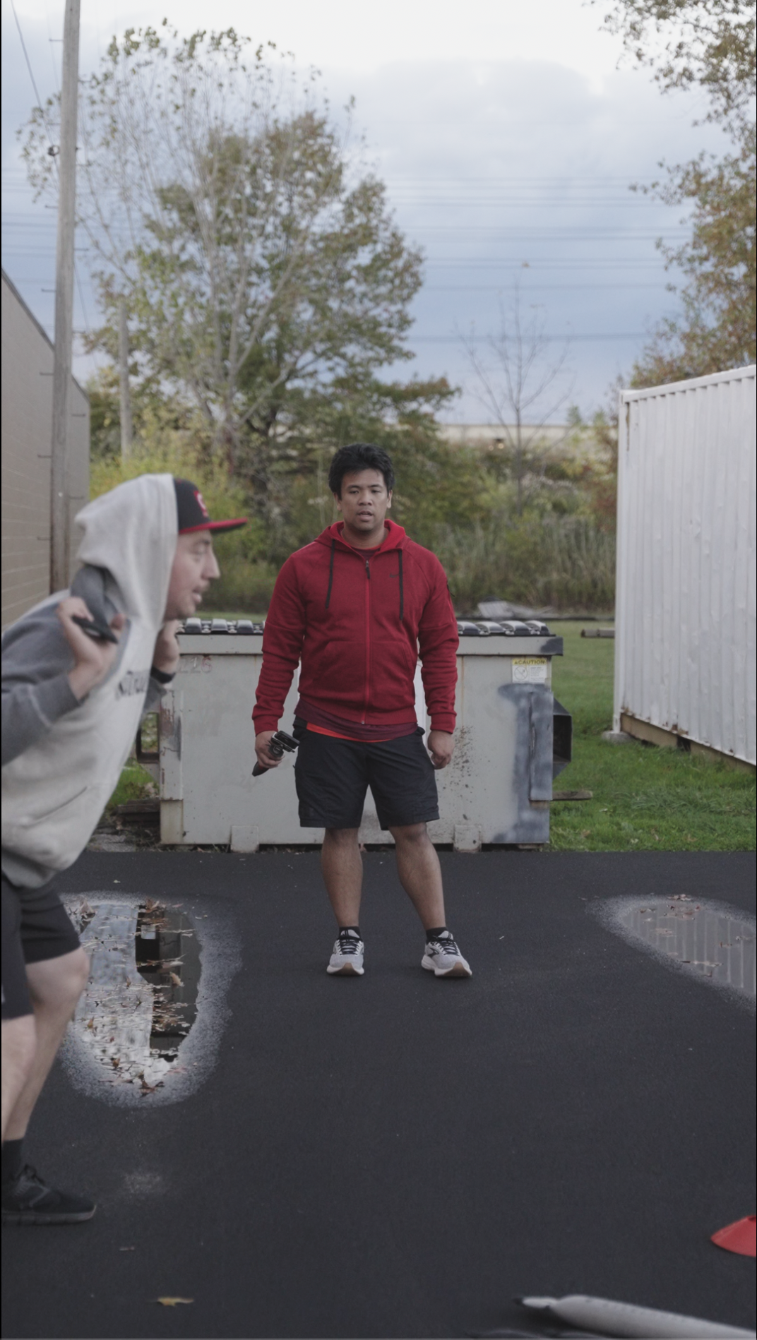 Two men, one in a hoodie and the other in a red jacket, are outdoors on a paved surface with puddles, trees, and a grey sky in the background.