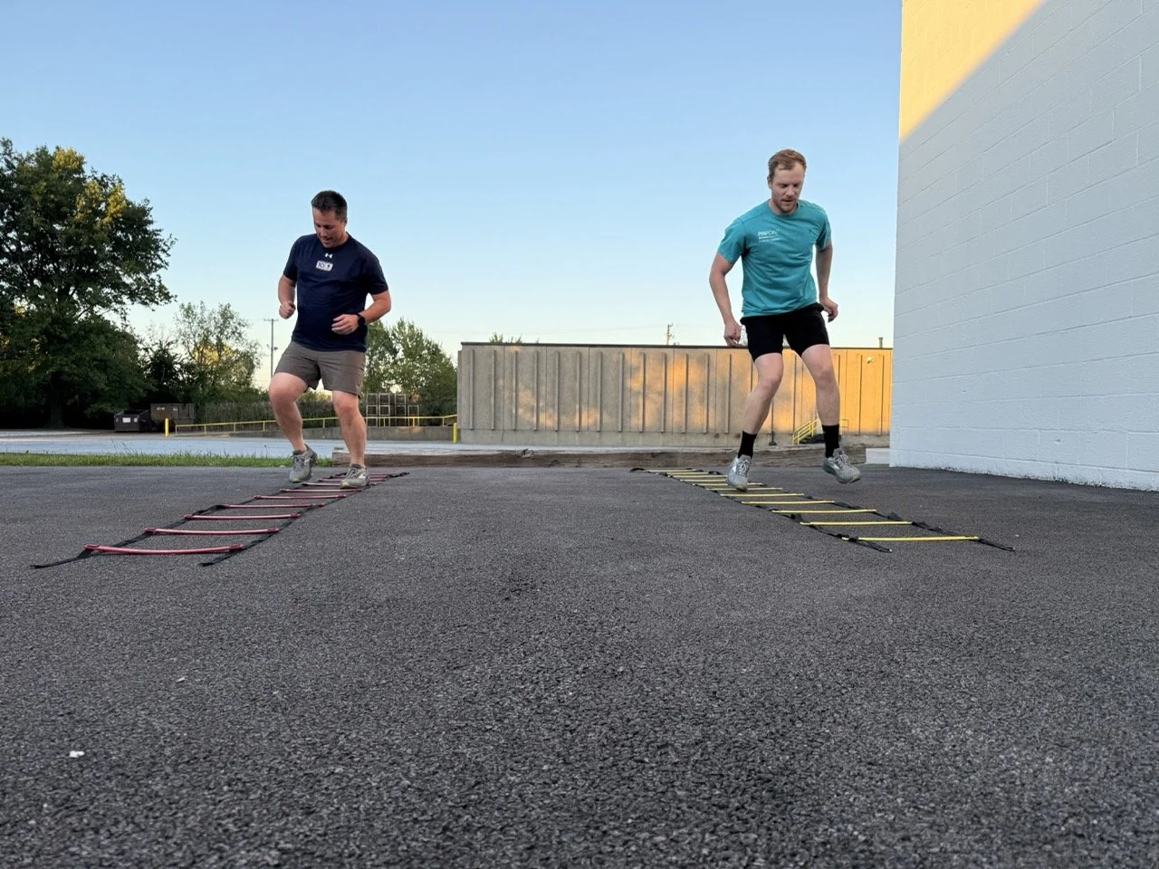 Two men training outdoors, jumping through agility ladders on a paved surface during sunset.