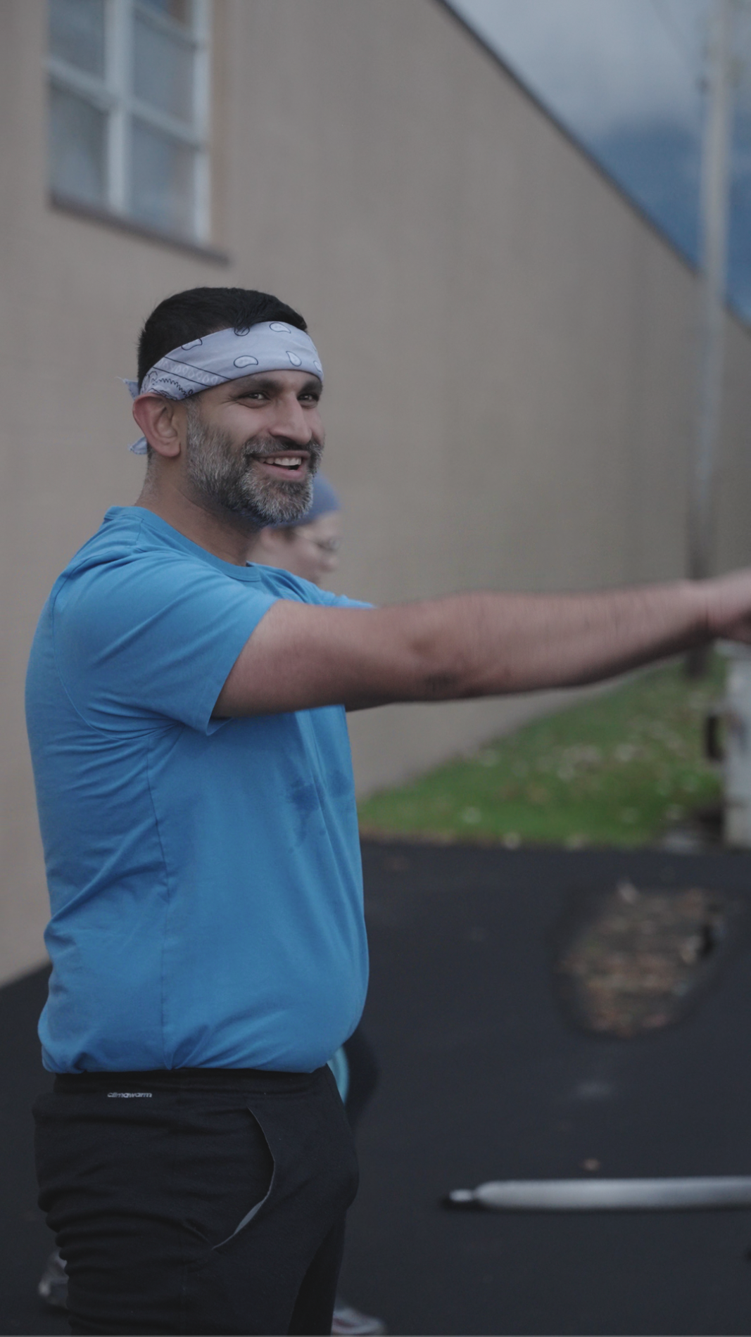 A man smiling, wearing a blue t-shirt and a light blue bandana, standing outdoors near a building with a window, with his arm extended forward.