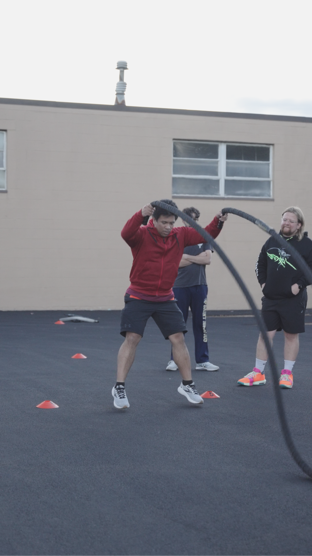 A young man in a red jacket and black shorts participating in battle rope training outdoors, with two other people standing nearby on a paved surface with orange cones marker.