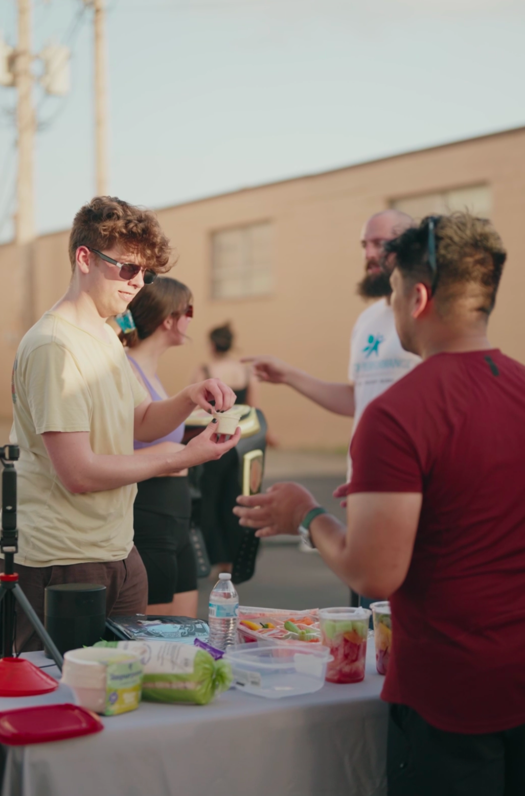 People at an outdoor event around a table with food and drinks, engaging in conversation.