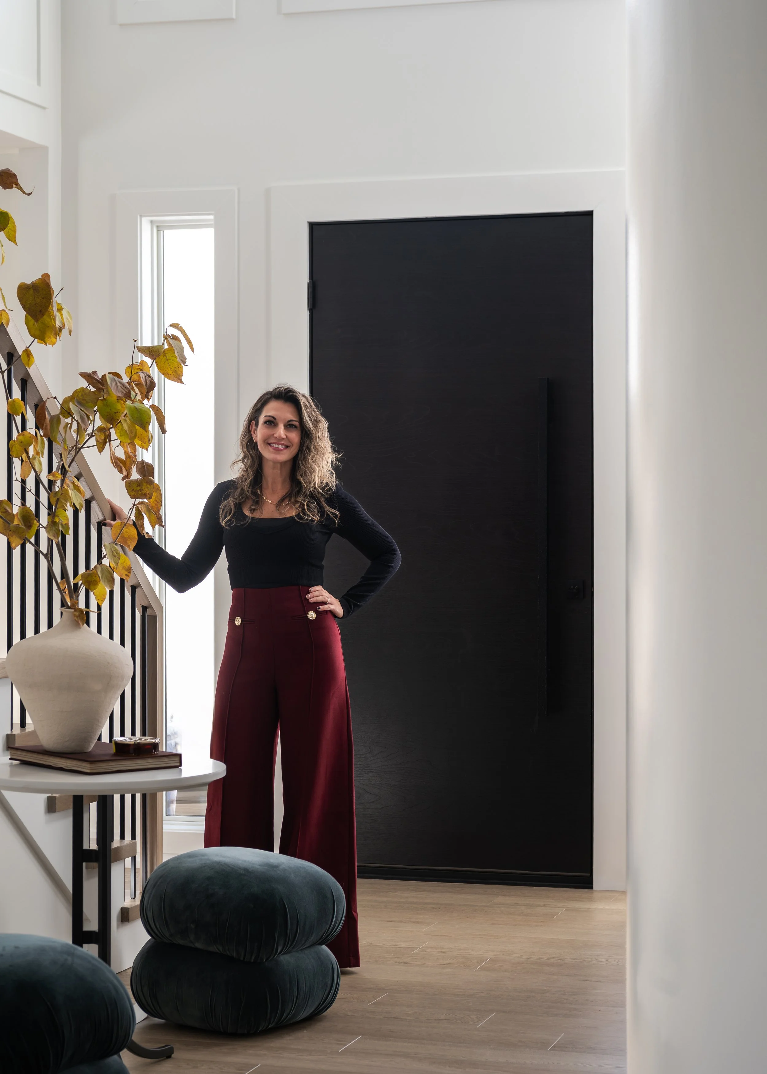 A woman with curly hair smiling and standing in a modern, well-lit interior near a black door and a staircase with a wooden railing. She is wearing a black long-sleeve top and high-waisted burgundy wide-leg pants.