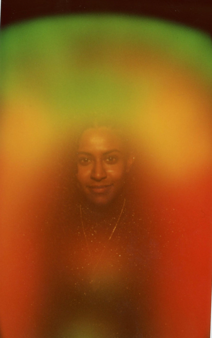 A woman with a gentle smile, wearing a dark, sparkly top and a gold necklace, with a colorful, blurry background.