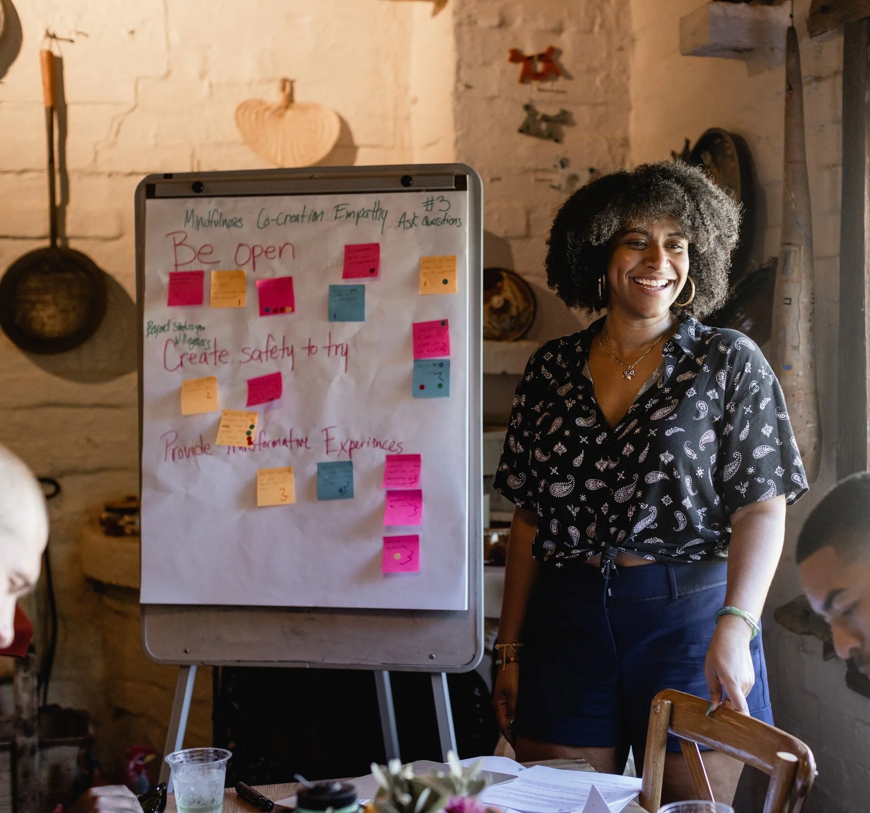 A woman with curly gray hair smiling in front of a whiteboard with notes and colorful sticky notes, standing in a room with wooden walls and various decorations.