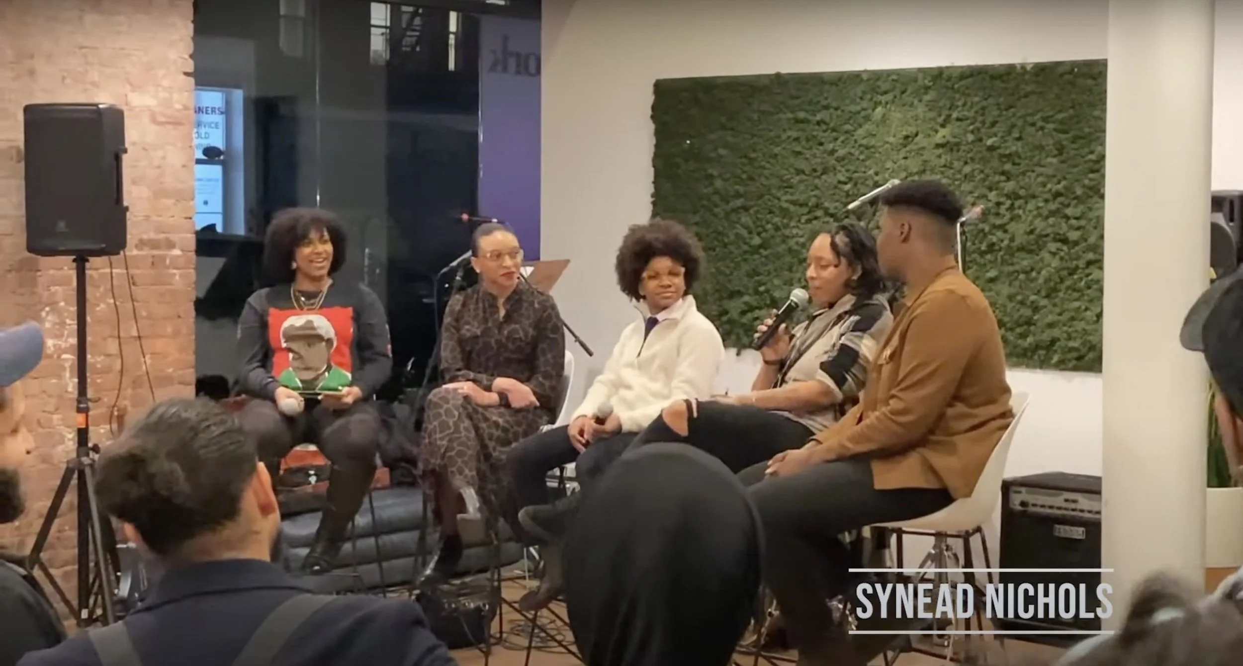 Panel discussion with five women seated on stage, one holding a microphone, in front of an audience, inside a room with brick and white walls and a green moss art piece.
