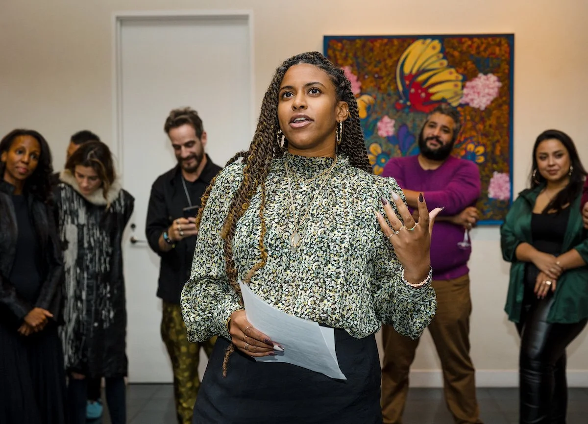 A young woman with braids speaking and gesturing, holding a piece of paper, in front of a group of diverse adults at an art gallery or event space, with a colorful floral art piece on the wall behind.