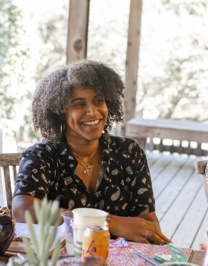 Smiling woman with curly gray hair wearing a black patterned shirt, sitting at a table outdoors, with a wooden deck and trees in the background.