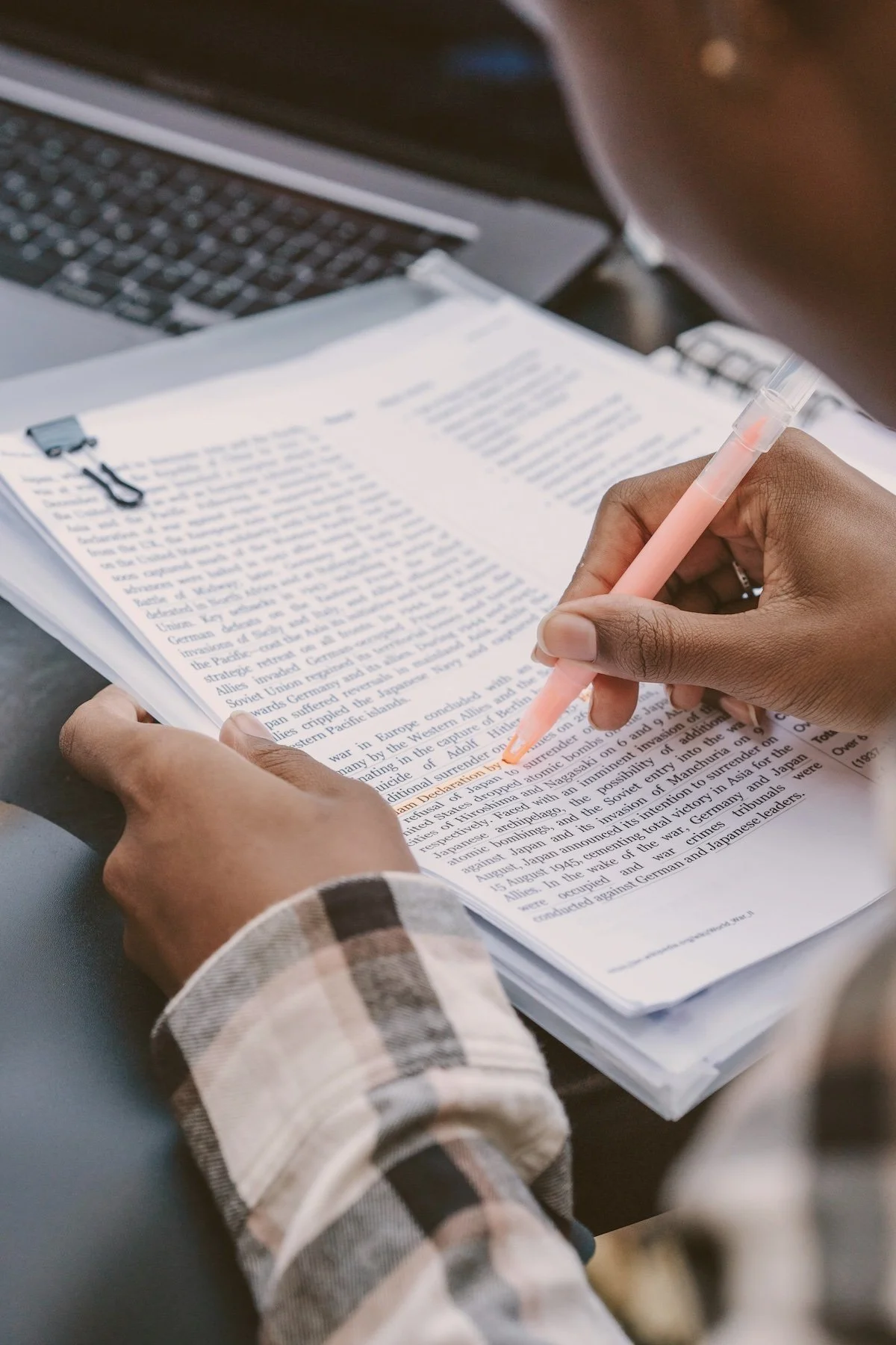 Person with a plaid shirt marking a text with a pink highlighter in a book, next to a laptop.