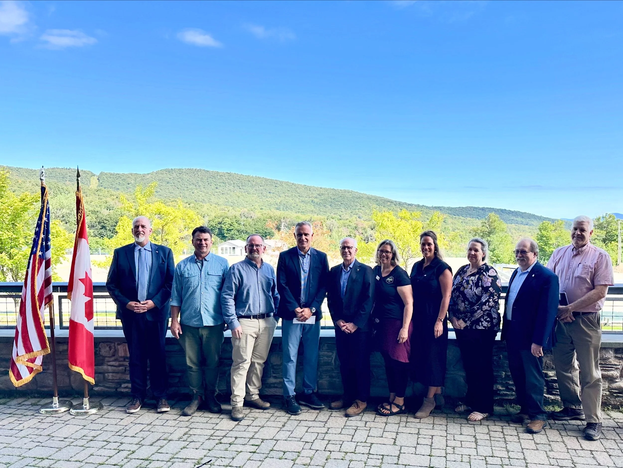 group of men and women in professional clothing stand next to each other. American flag and canadian flag are next to them with a mountain view in the background.