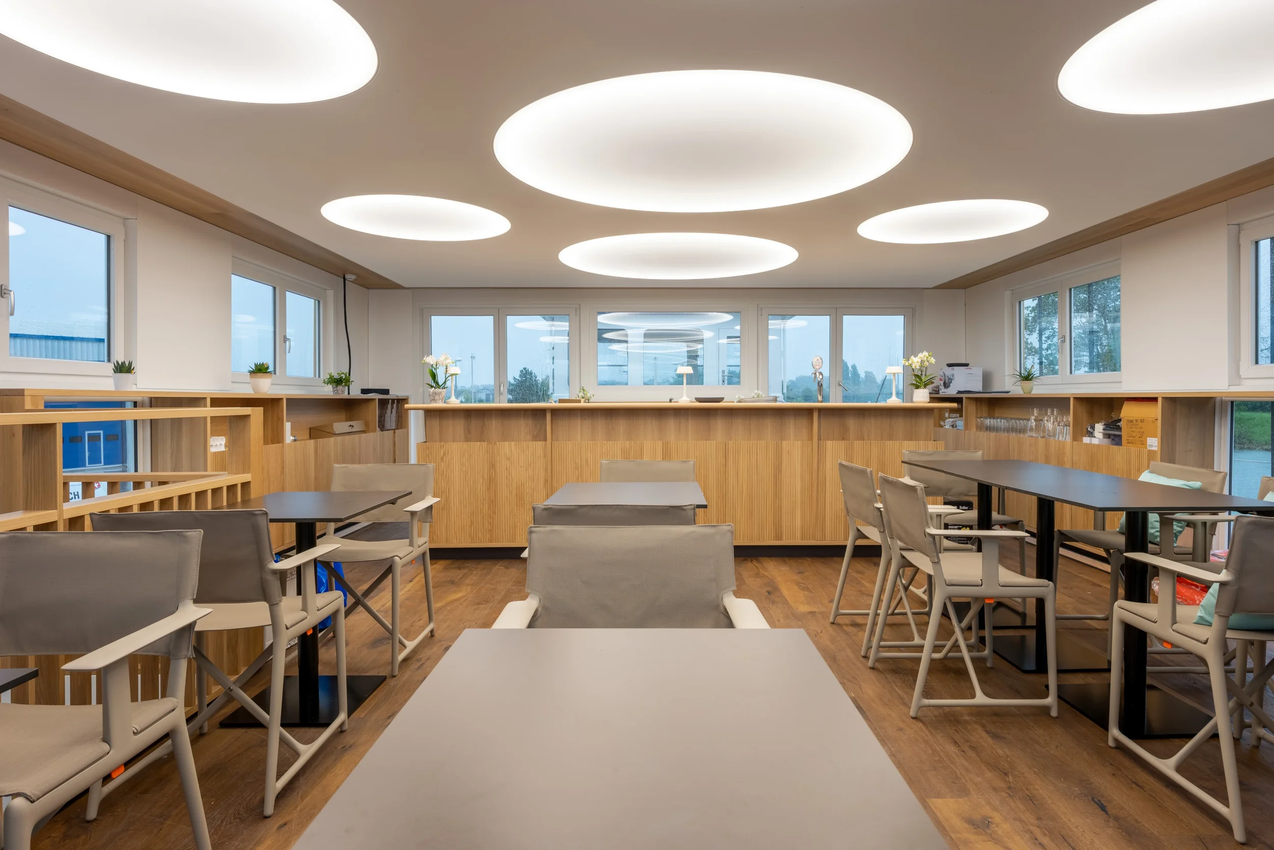 Gemütlicher Essbereich mit großen Fenstern, weißen Wänden, Holzböden, modernen runden Deckenleuchten, mehreren Tischen und Stühlen, einigen Pflanzen sowie einer Küchen- oder Servicetheke im Hintergrund.