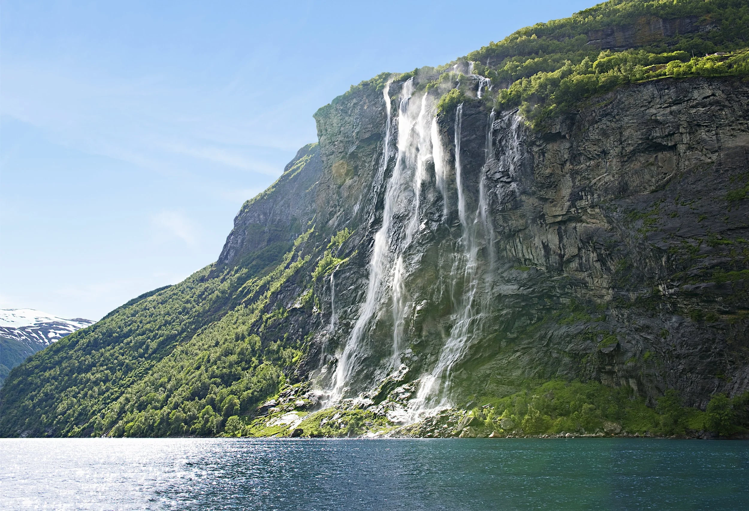 Ein Wasserfall, der an einer Klippe in einem Fjord oder See in Norwegen herabstürzt, umgeben von grünen Bergen.