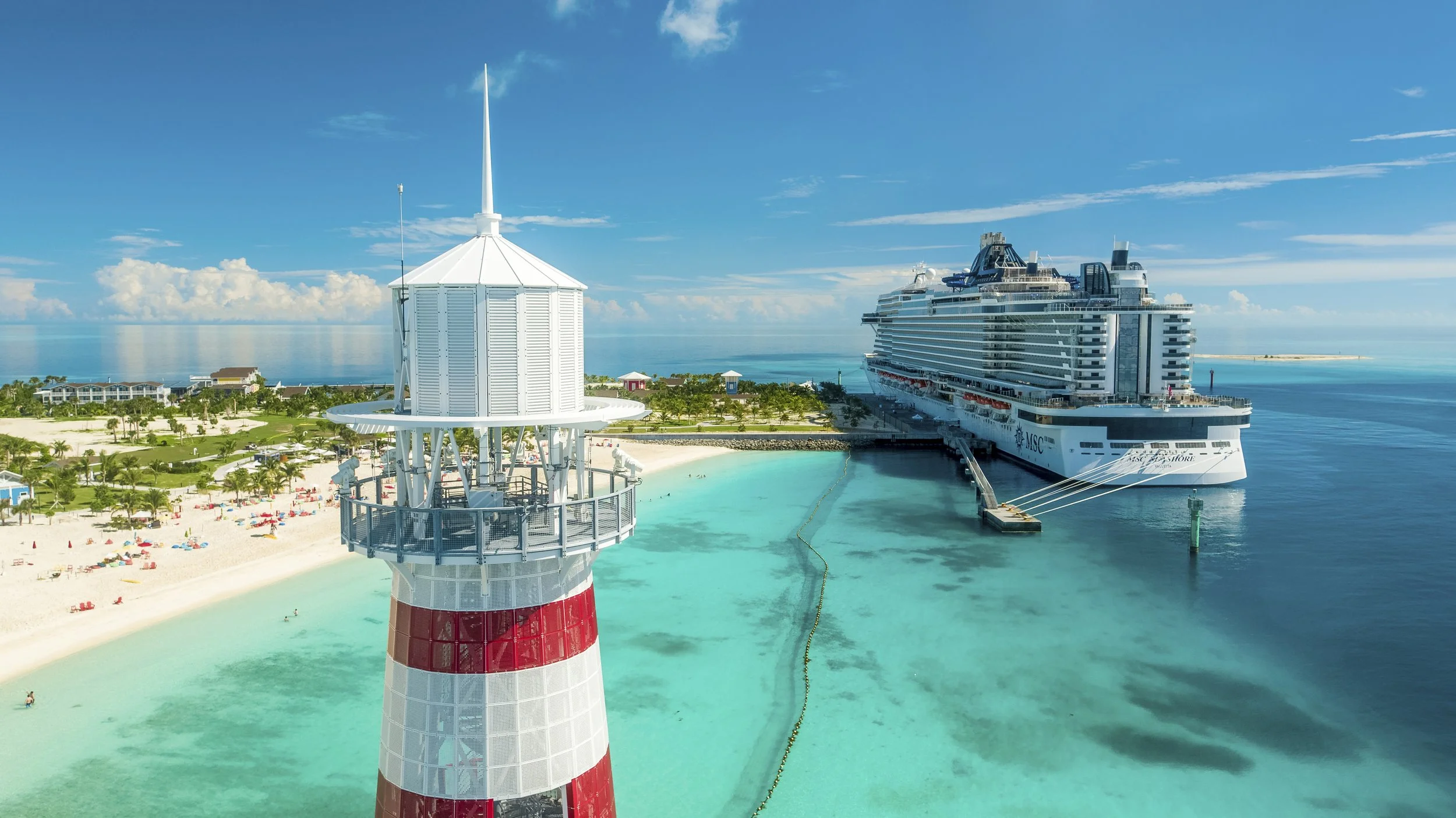 Ein Leuchtturm am Strand mit einem Kreuzfahrtschiff im Hafen und blauem Himmel im Hintergrund.