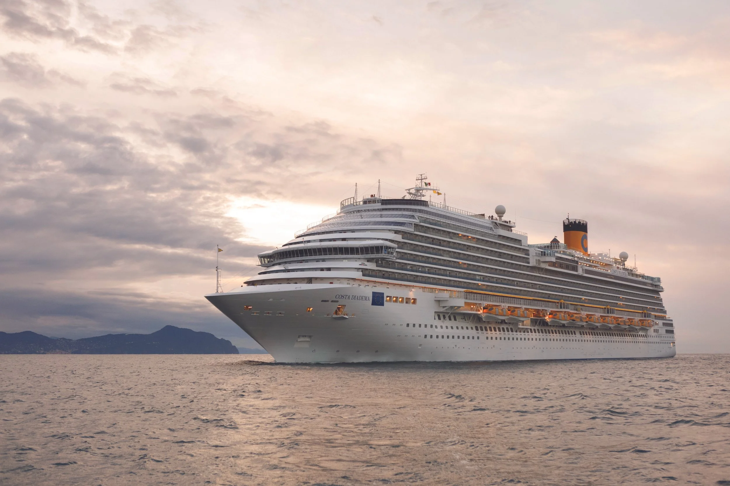 Ein großes Kreuzfahrtschiff namens Costa Diadema auf dem Wasser, bei Sonnenuntergang oder Dämmerung, mit Wolken am Himmel und Bergen im Hintergrund.
