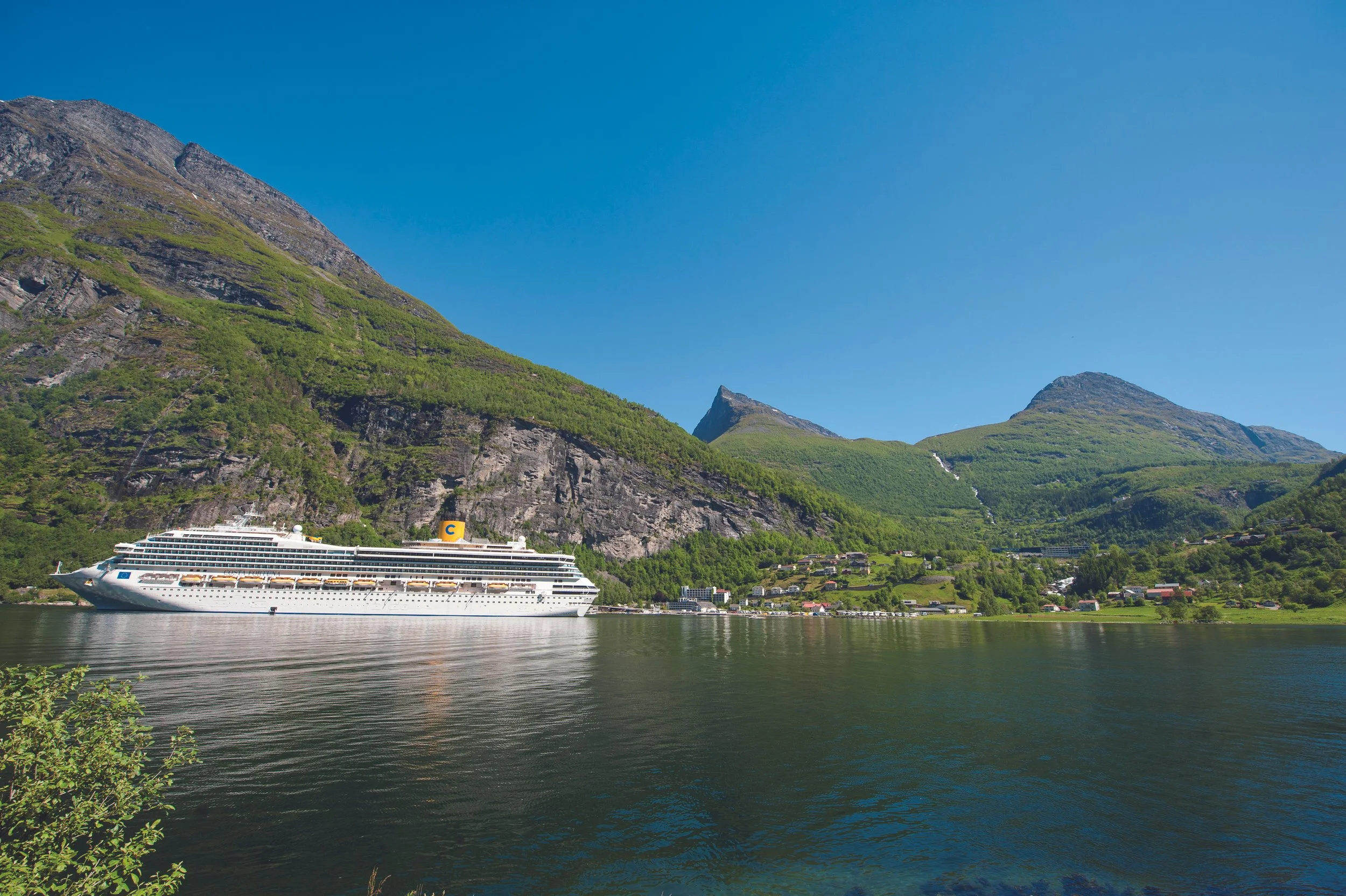 Ein Kreuzfahrtschiff auf einem Fjord vor grünen, steilen Bergen unter einem klaren blauen Himmel.