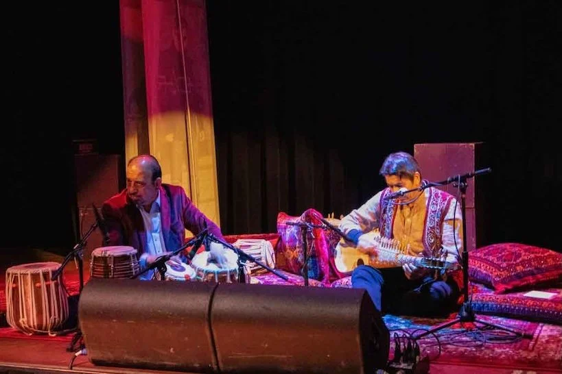 Two musicians on stage - one playing a string instrument, and the other playing hand drums.