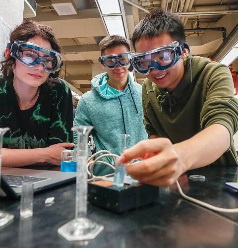 Three people wearing safety goggles and handling test tubes at a table. 