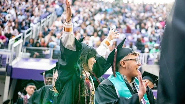 Two graduates in college graduation robes, smiling with a large crowd in bleachers behind them.