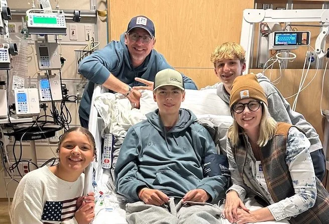 Two adult and two children smiling and standing around a hospital bed with a teenager sitting on it.