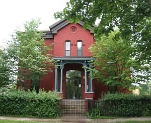 The exterior of a red brick house with green trees on hedges in front of it. 